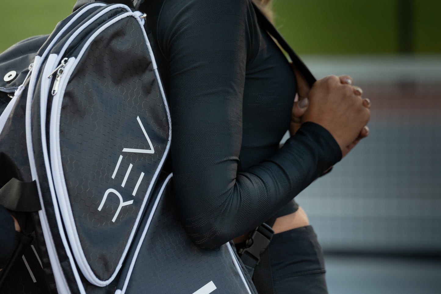 A person in black athletic wear carries the PRO Player Pickleball Bag by REV Pickleball, featuring bold white REV lettering. The blurred tennis net and green court suggest a pickleball setting—ideal for storing gear or insulated paddles.
