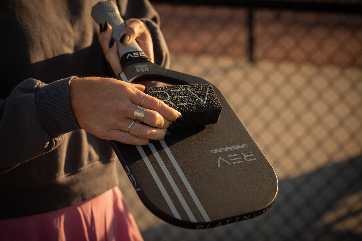 A person cleans a black raw carbon fiber paddle labeled "JAY PLATINUM" with the REV Pickleball Carbon Fiber Paddle Cleaner; they wear a sweatshirt and rings. Sunlight bathes a blurred chain-link fence in the background.