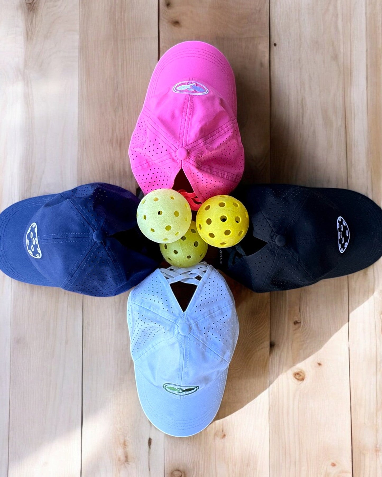 Four Fresh Pickle Pickleball Caps—pink, black, blue, and white—form a cross on a wooden floor with three yellow pickleballs in the center, showcasing perfect style and function for your next game.