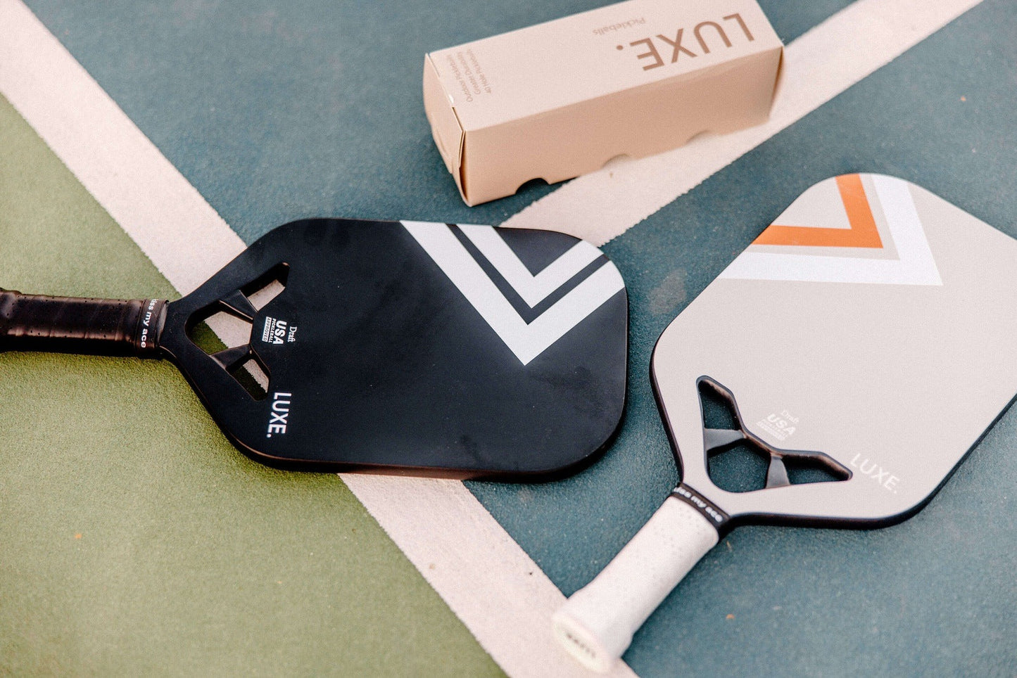Two LUXE Pickleball Draft paddles, one featuring gray chevrons, rest on a worn green court near a white line. Above them is a beige Luxe paddle box. Both paddles are USA Pickleball Approved.