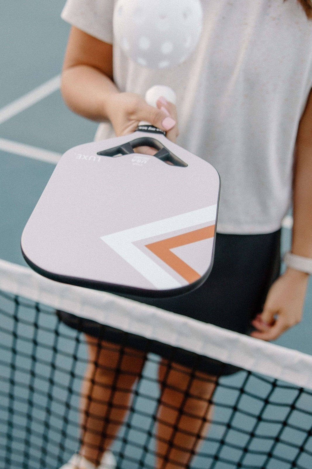 A person in a white shirt and black skirt stands at a pickleball net, holding out the LUXE Pickleball Draft carbon fiber paddle and a perforated white ball; only their torso and arm are visible, with the court surface in the background.