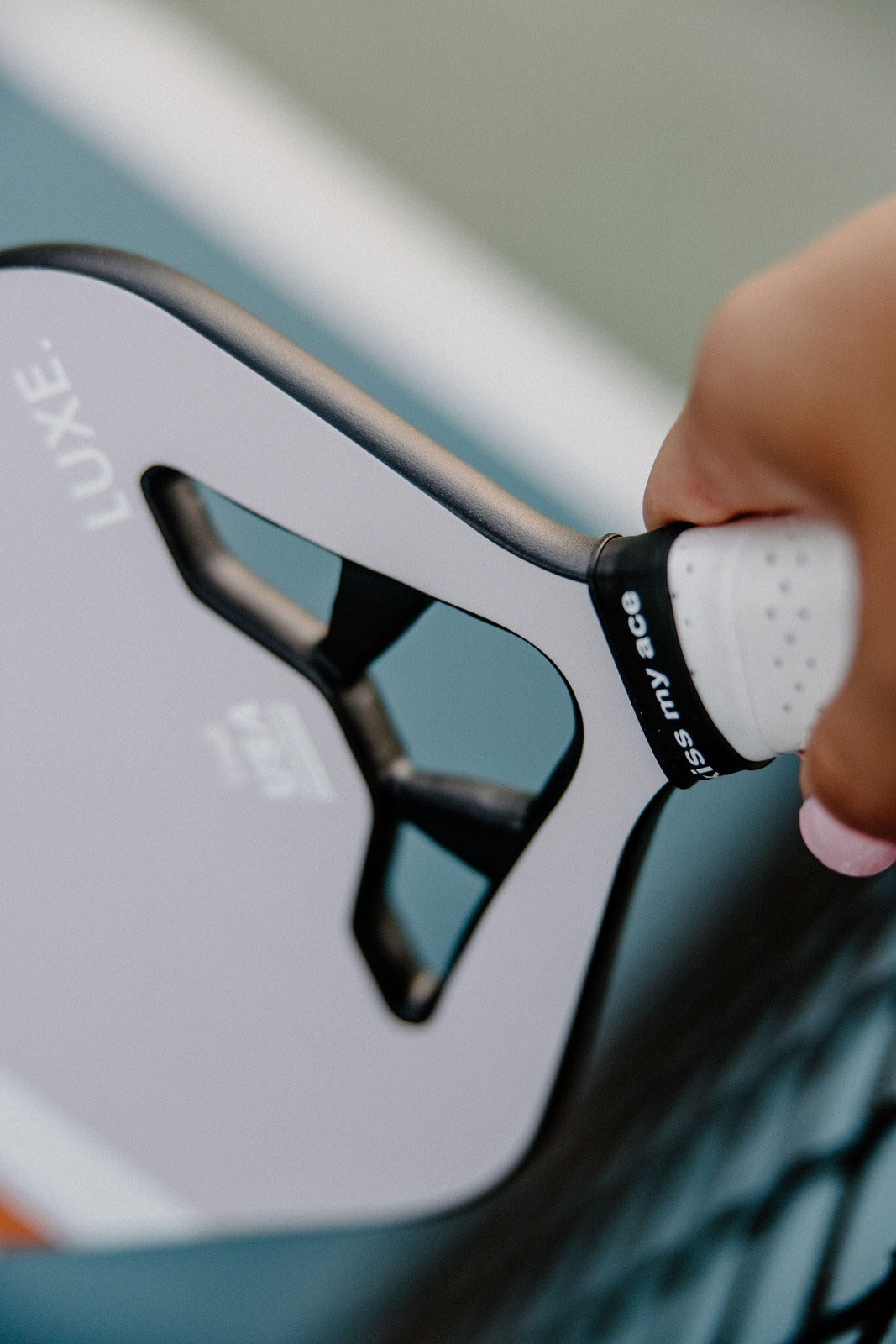 A close-up shows a hand gripping the white handle of the LUXE Pickleball Draft paddle, USA Pickleball Approved, with the brand name visible above a blurred blue and green court. The paddle's edge and part of the hand are in sharp focus.