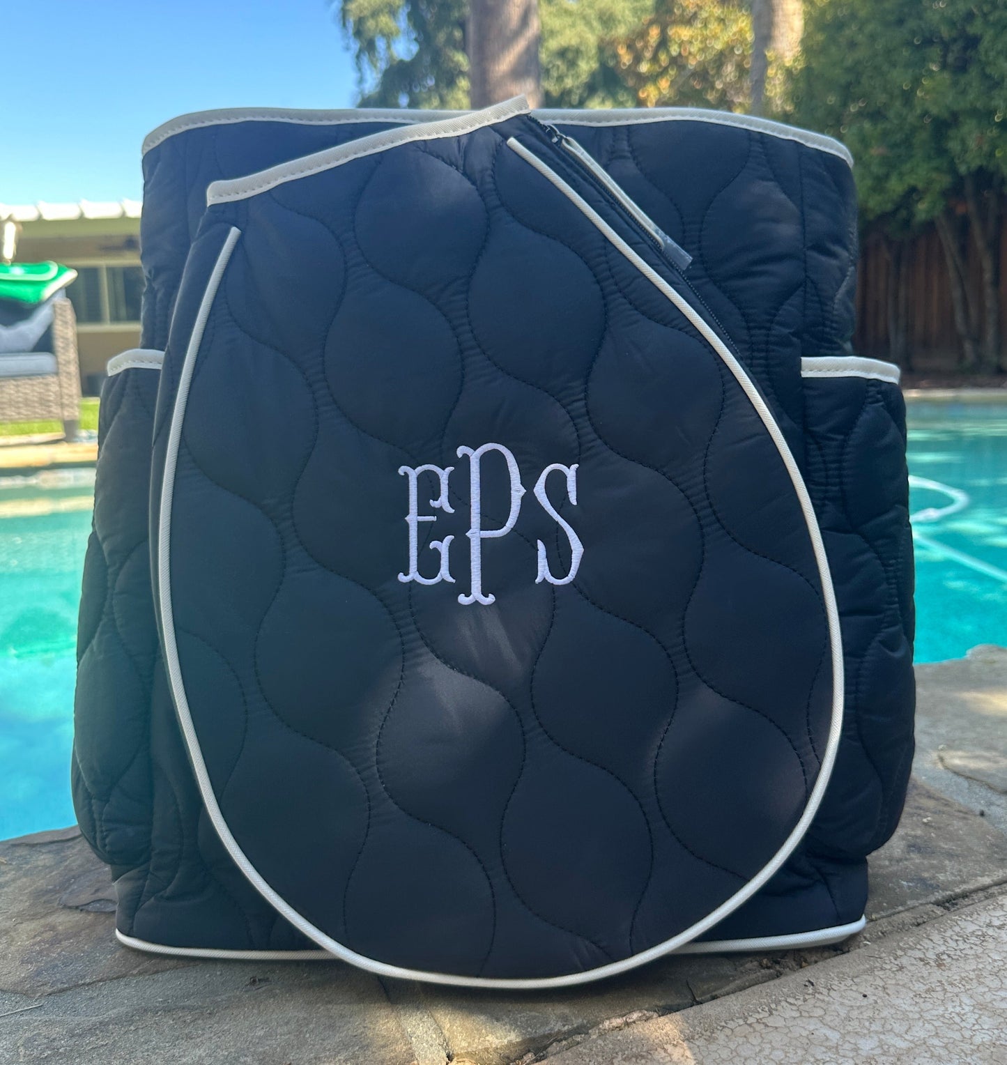 A Park Ave Tennis Convertible Bag/Backpack by Queen of the Court in navy blue with white piping and EPS monogram rests on a stone ledge by a pool, with trees, a wooden fence, and part of a house visible in the sunny background.