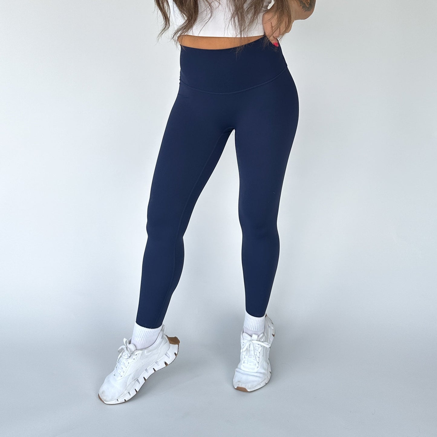 Against a plain white background, a person (head not visible) wears DYM Athletics Flex & Flow Legging in navy blue, featuring a high-waisted seamless fit and buttery soft fabric, paired with a cropped white top, socks, and chunky white sneakers.