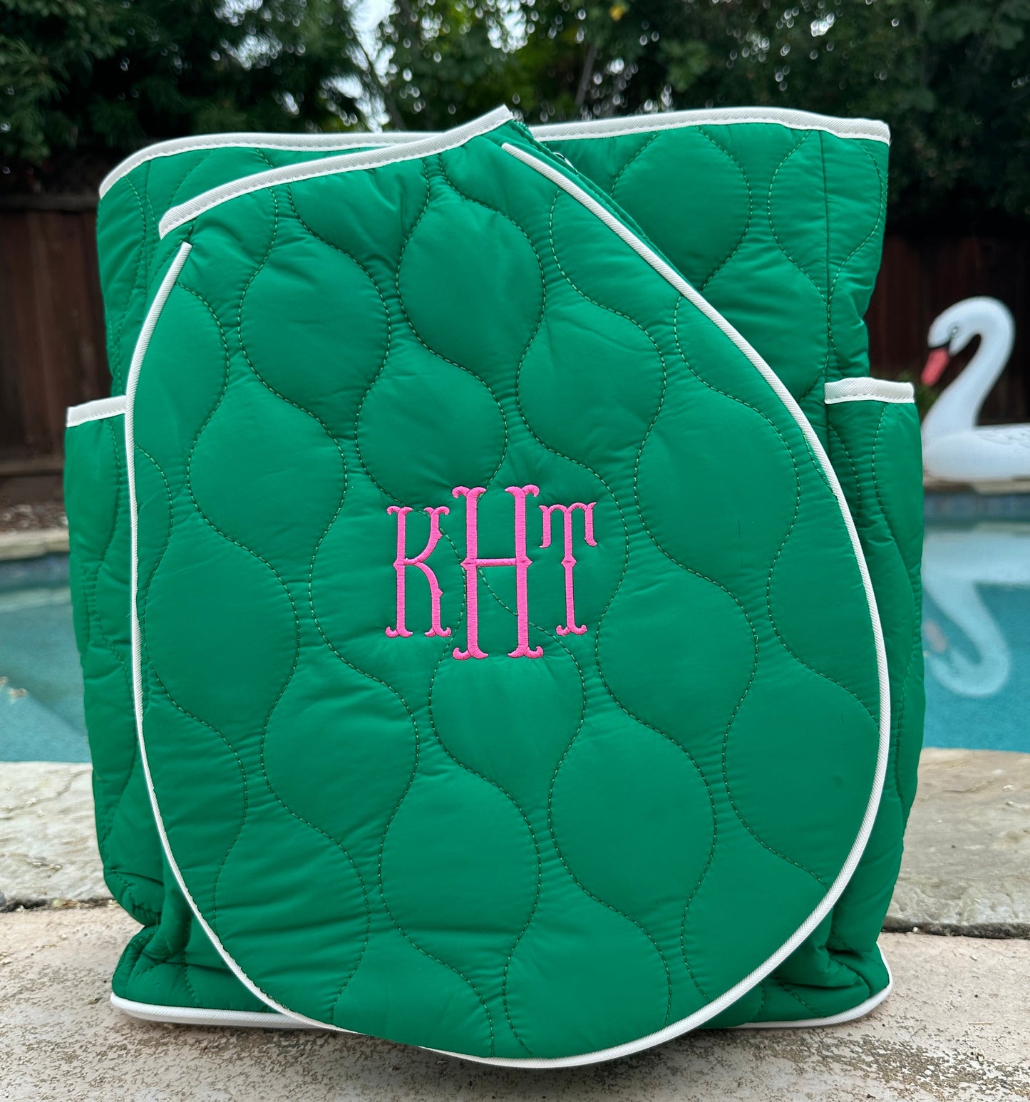 The Queen of the Court Park Ave Tennis Convertible Bag/Backpack in green with white trim and pink monogrammed "KHT" sits poolside, featuring side pockets and a curved front flap beside a swan float, blue water, and greenery.