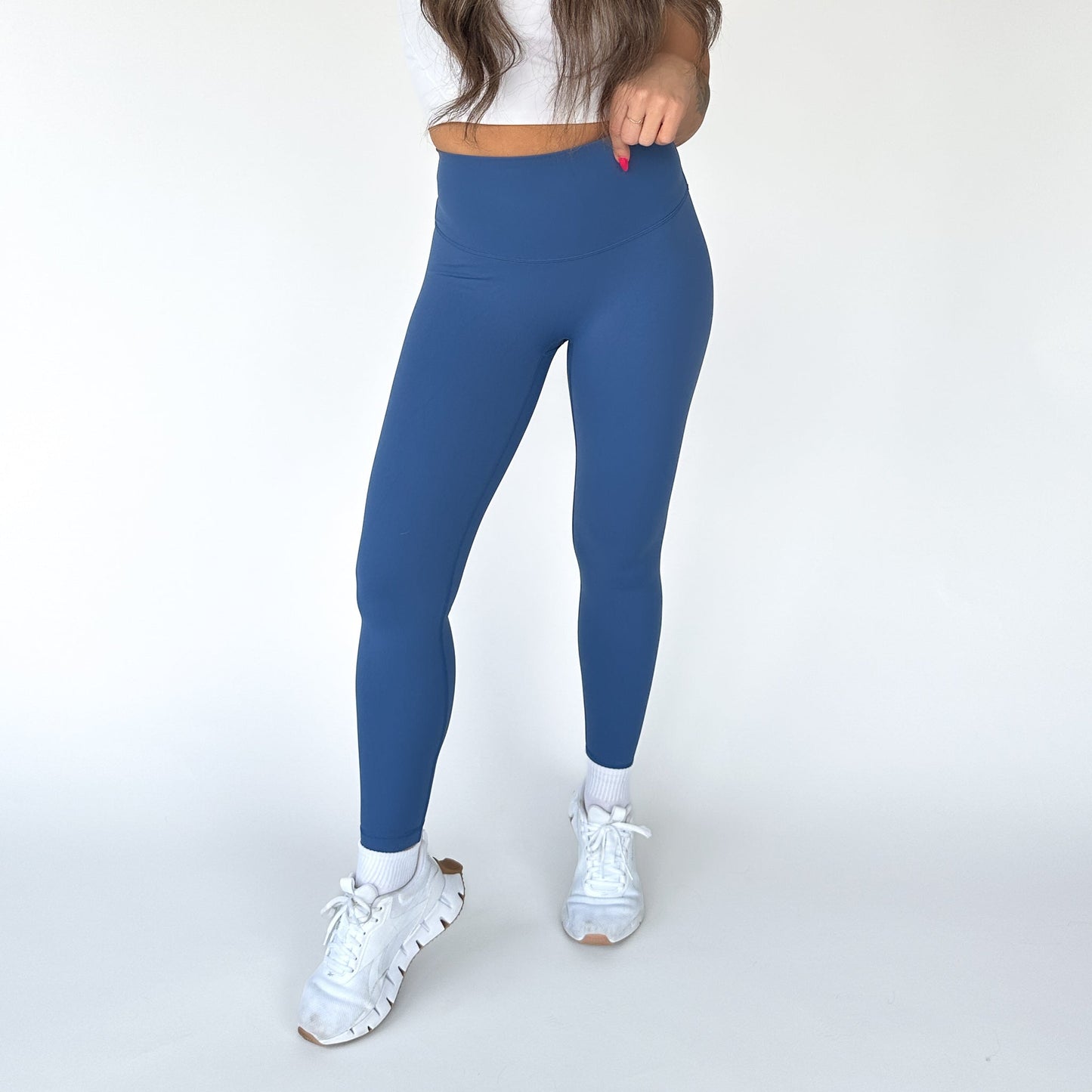 A woman poses against a white background wearing DYM Athletics Flex & Flow Legging in high-waisted blue, paired with a white cropped top, white ankle socks, and sneakers. Her long brown hair falls over her shoulder as she holds a strand.