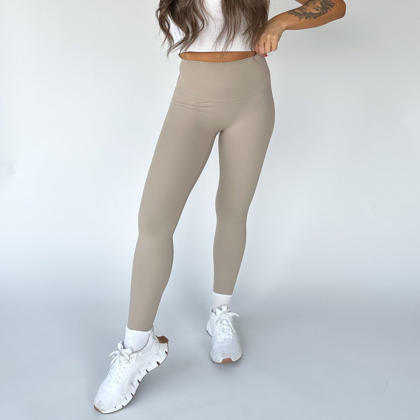 A woman poses against a white background wearing DYM Athletics’ Flex & Flow Legging—high-waisted and buttery soft—paired with a cropped white top, crew socks, and chunky sneakers. Her tattooed left arm is bent; her face is not visible.