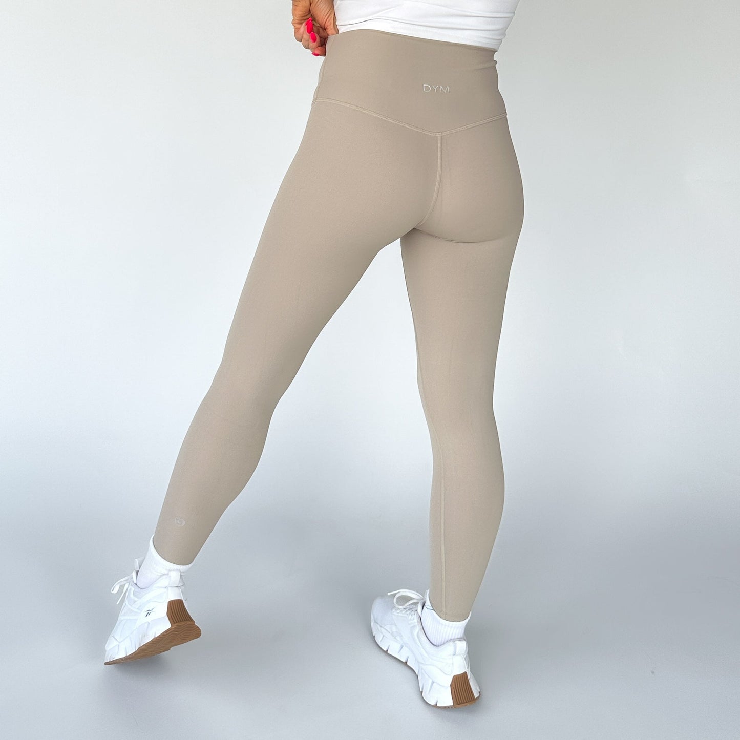 A person stands away from the camera in DYM Athletics Flex & Flow Leggings, beige high-waisted and seamless with a subtle waistband logo, paired with a white top and sneakers. The background is plain light gray; one hand rests on their hip.