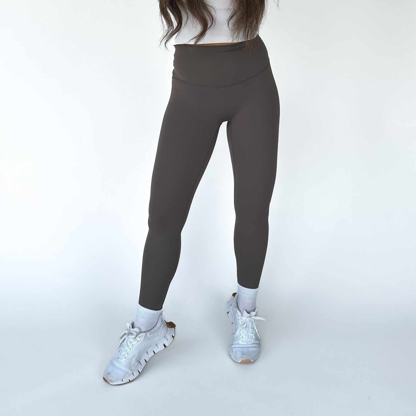 Wearing DYM Athletics Flex & Flow Legging in high-waisted dark gray, made from buttery soft fabric, paired with a white top, socks, and sneakers. Standing casually against a plain white background with feet slightly apart.