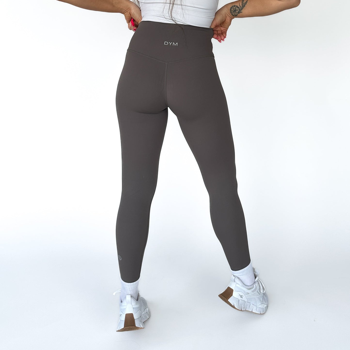 A person stands against a white background, facing away, wearing high-waisted dark gray Flex & Flow Leggings by DYM Athletics with “DYM” on the waistband. Their hands are on their hips, and part of a tattoo is visible on their left arm.