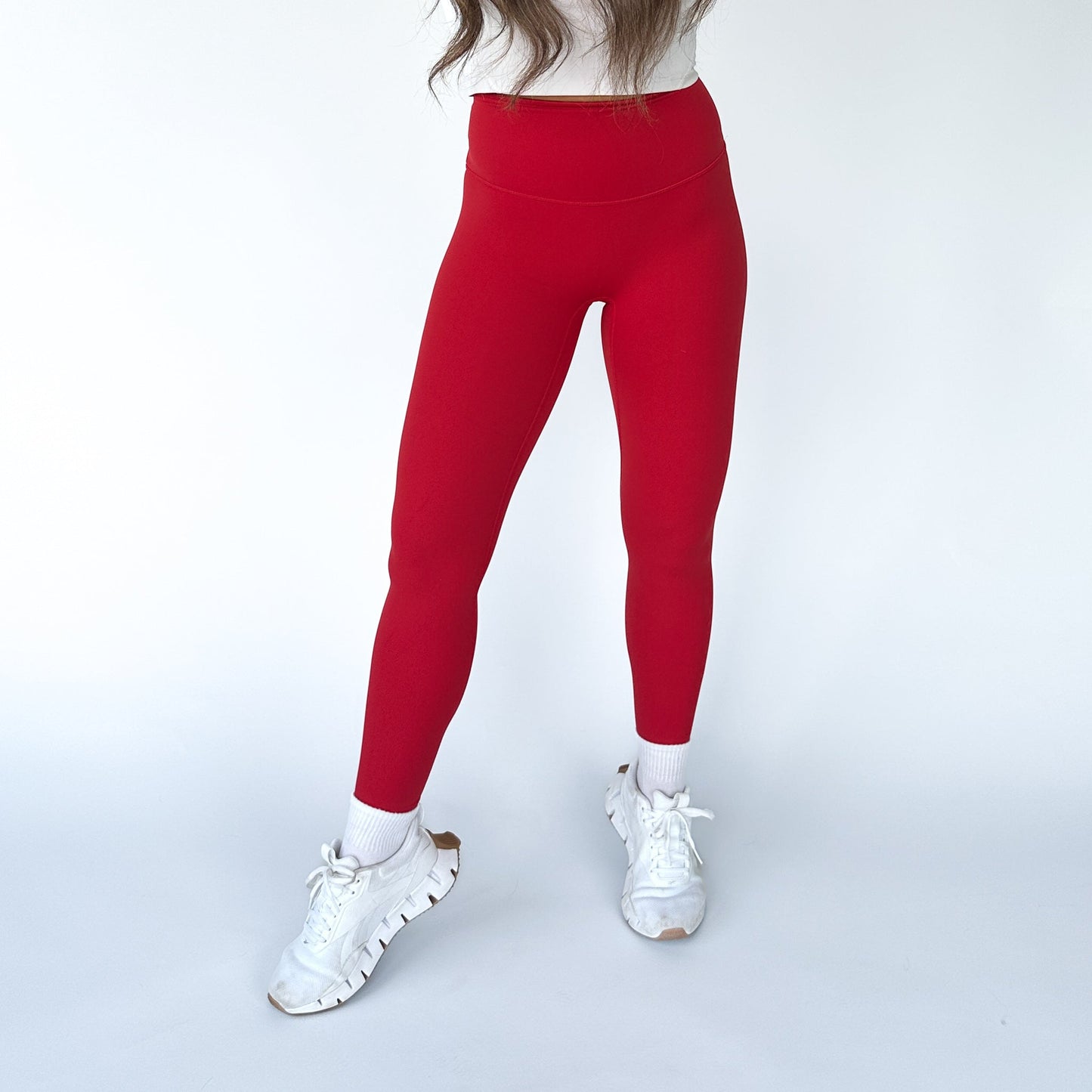 Against a plain white background, only the lower half of a person is shown sporting DYM Athletics’ Flex & Flow Legging in bright red, paired with white socks and athletic shoes—highlighting the stylish, buttery soft activewear.