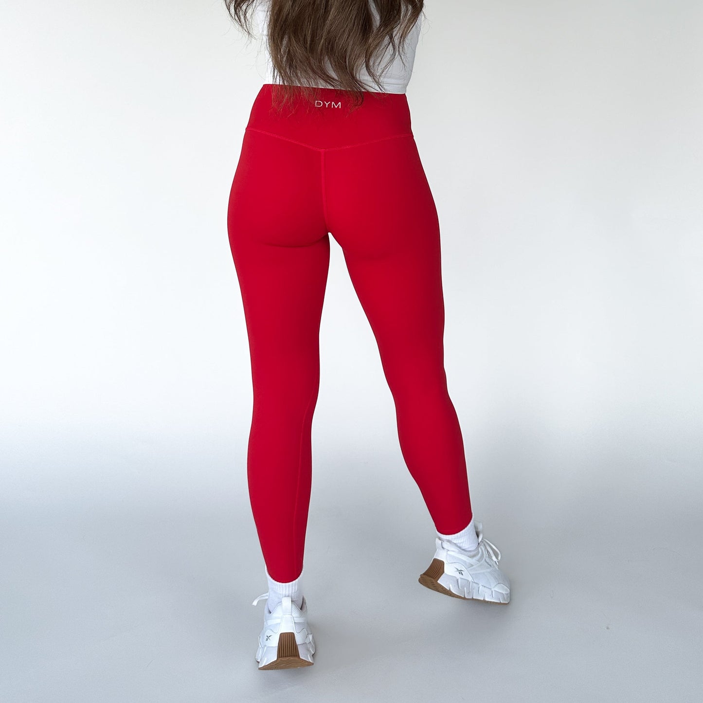 A person with long brown hair faces away, wearing DYM Athletics’ Flex & Flow Legging in bright red, a white long-sleeve top, and white sneakers with brown soles. The minimal studio setting highlights the bold DYM activewear.