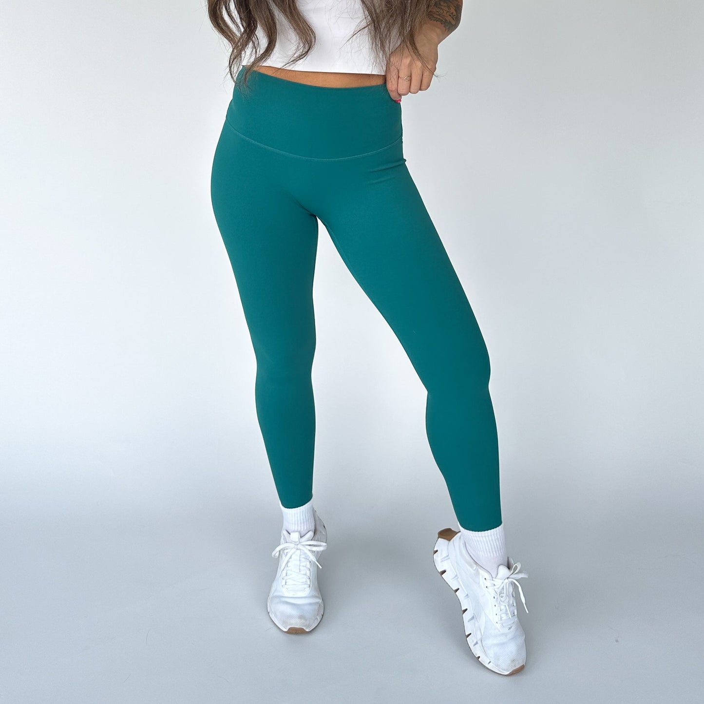 A person stands against a plain white background wearing DYM Athletics' Flex & Flow Legging in teal with a white crop top, socks, and sneakers. Their long hair is partially visible, and they strike a relaxed pose with one leg slightly bent.