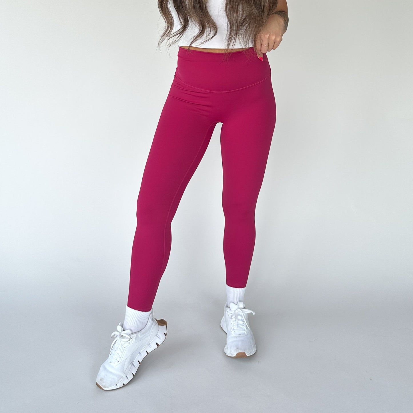 A person with long brown hair wears DYM Athletics' Flex & Flow Legging in magenta, styled high-waisted with a white crop top, crew socks, and chunky sneakers. The seamless leggings feature buttery soft fabric. Their head is out of frame.