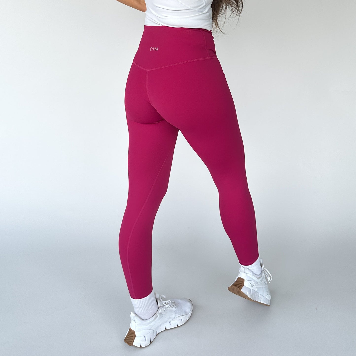 Wearing DYM Athletics' Flex & Flow Legging in bright pink, paired with a white top, socks, and chunky sneakers, a person poses from behind against a white background to showcase the leggings' fit, seam details, and buttery soft fabric.