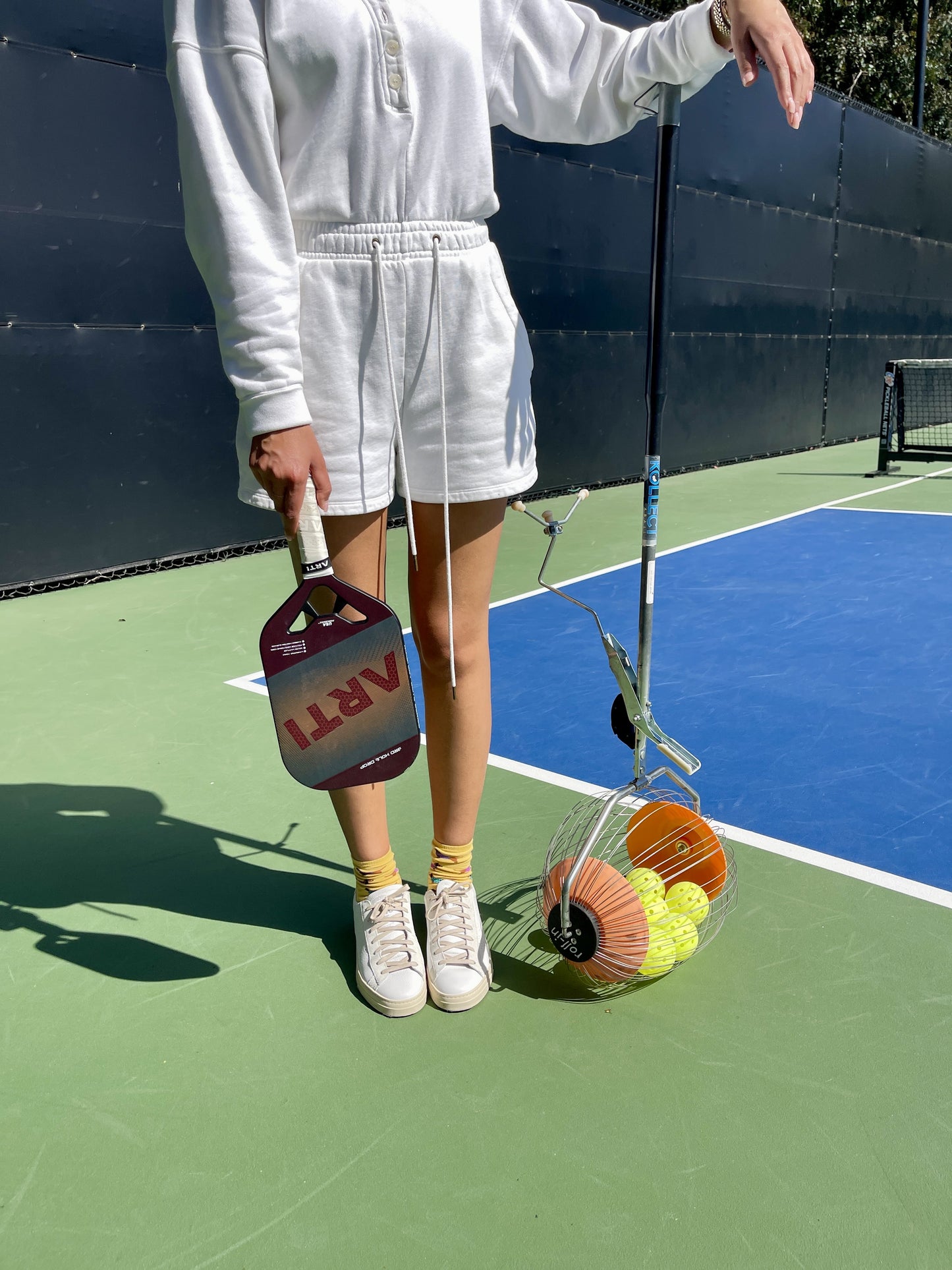 Wearing a white hoodie and shorts, someone stands on a tennis court holding the ARTI Carbon Fiber "MASTERY ELITE 1.0" paddle by ARTI. Their other arm rests on a basket of yellow pickleballs and two orange cones; only their body is visible.