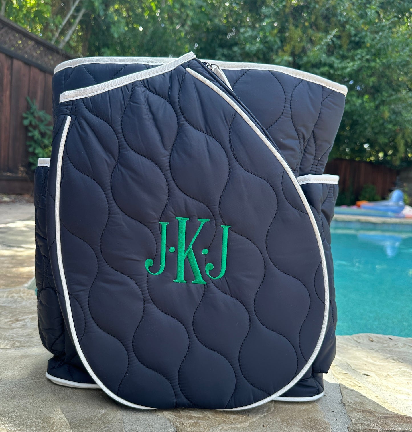 The Park Ave Tennis Convertible Bag/Backpack by Queen of the Court, in navy blue with white trim and monogrammed green initials JKJ, sits on a stone patio near a pool, surrounded by trees, a wooden fence, and yard furniture.