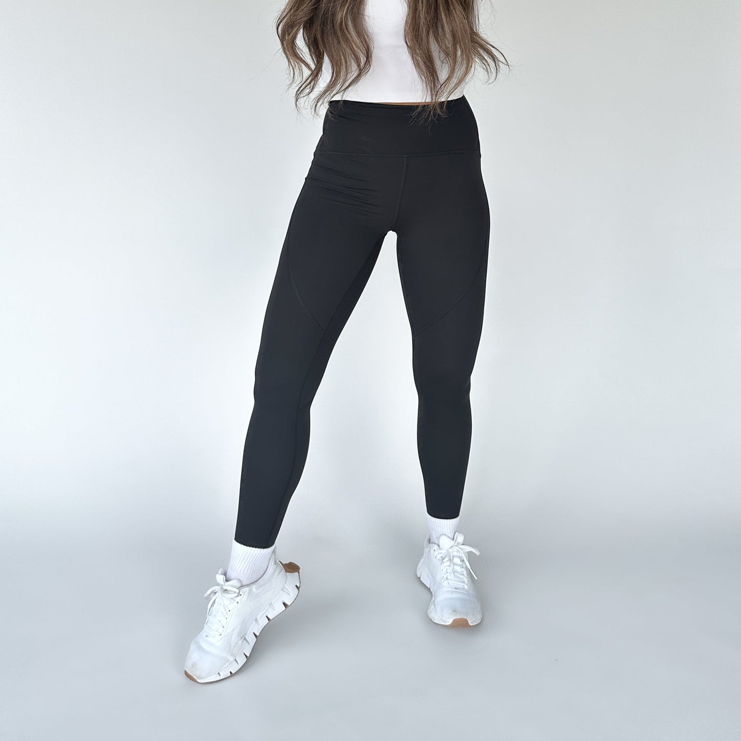 A person stands against a light background, wearing DYM Athletics’ Trainer Muscle Tone Legging with a white fitted top, white socks, and chunky white sneakers. Their face is hidden, and long brown hair falls over their shoulders.