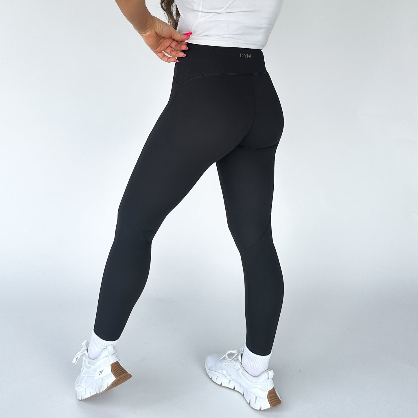 A person stands with their back to the camera against a plain white background, wearing a white t-shirt, DYM Athletics Trainer Muscle Tone Legging high-waist leggings, white athletic shoes with brown soles, and white ankle socks. Their left hand rests on their hip.