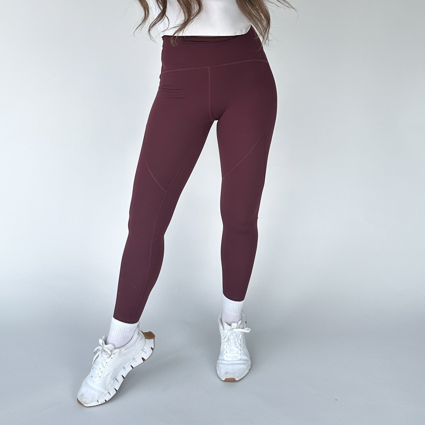 A person wears burgundy DYM Athletics Trainer Muscle Tone Legging, a white long-sleeve top, white socks, and chunky white sneakers against a plain light gray background. Their face is not visible.