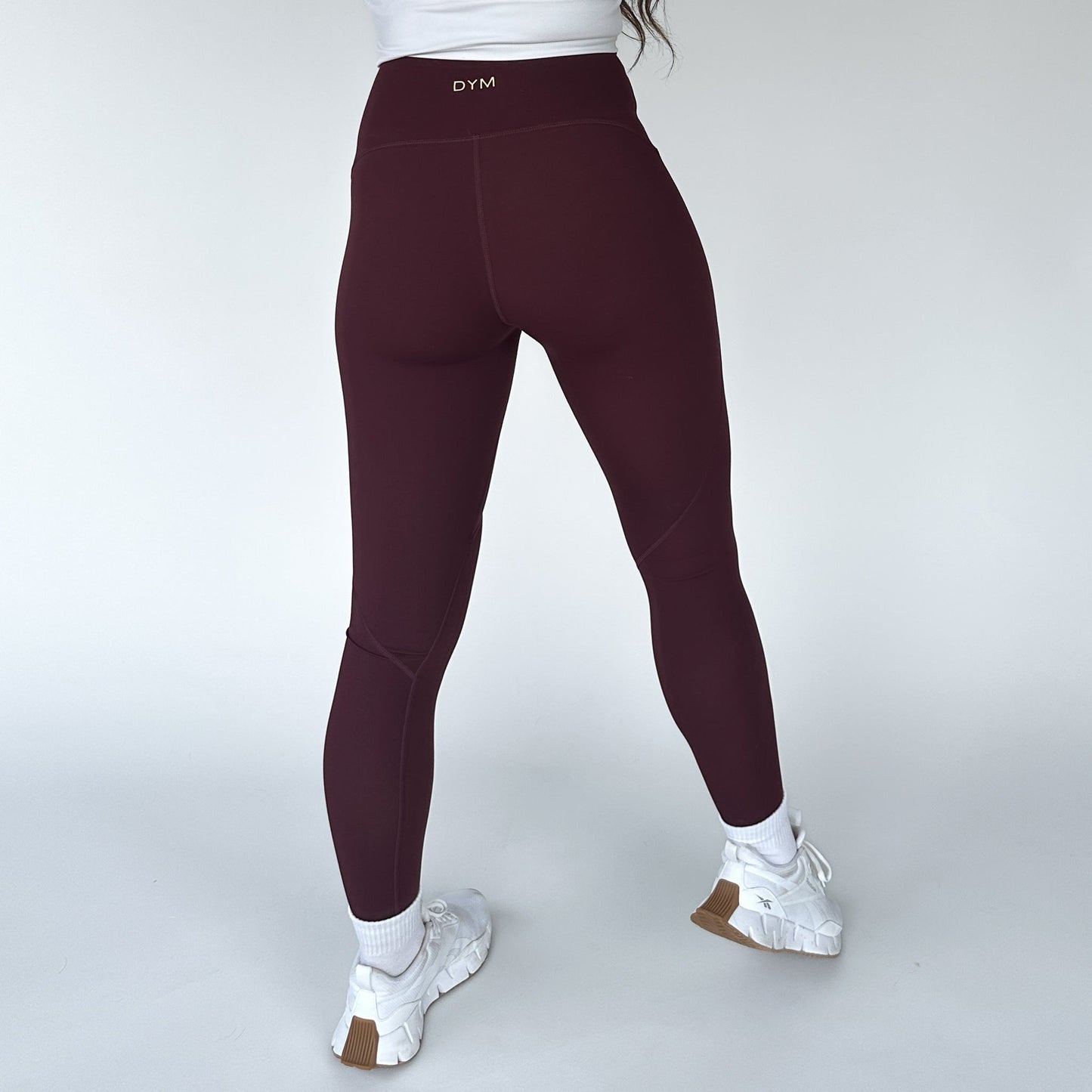 A person faces away, wearing DYM Athletics Trainer Muscle Tone Legging in burgundy with “DYM” on the waistband, a white top, socks, and chunky sneakers. The plain background highlights these stylish compression workout leggings.