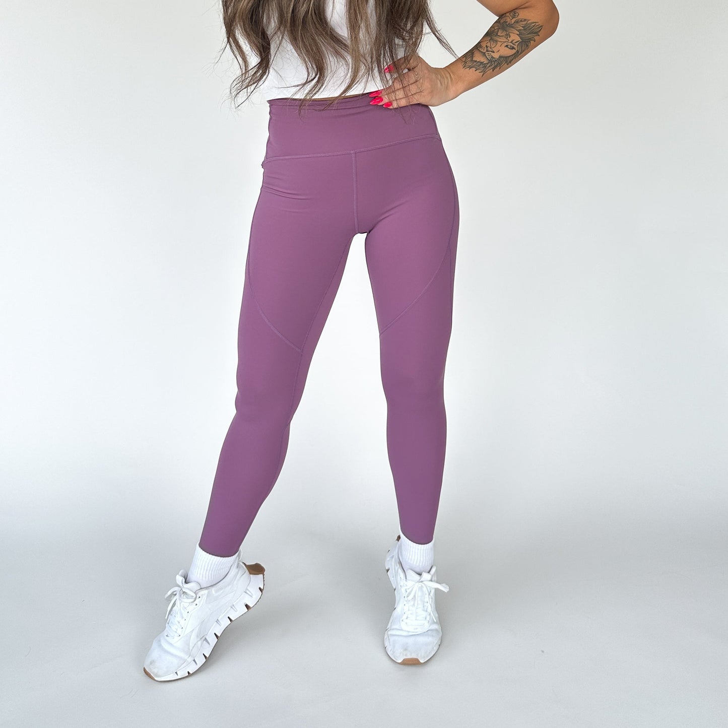 A person poses against a white background in DYM Athletics Trainer Muscle Tone Leggings (mauve), a white top, and chunky sneakers. Their long brown hair is loose, one hand on their hip, with a tattoo visible on their left forearm.