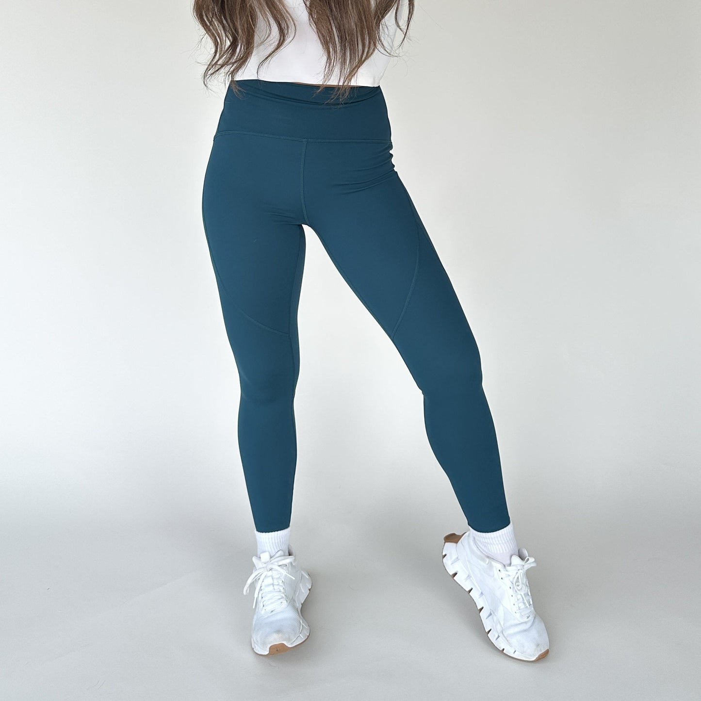 A woman stands against a white background wearing DYM Athletics Trainer Muscle Tone Legging in teal, a white top, and sneakers. Her long brown hair is visible but her face is out of frame, and one knee is slightly bent.