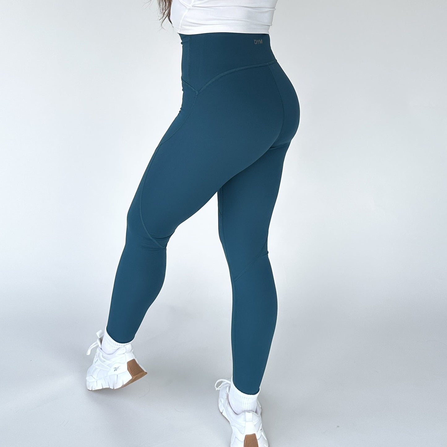 A person is pictured from behind in DYM Athletics Trainer Muscle Tone Legging, paired with a white short-sleeve top and white sneakers, set against a plain background to showcase the fit of these high-waist leggings.