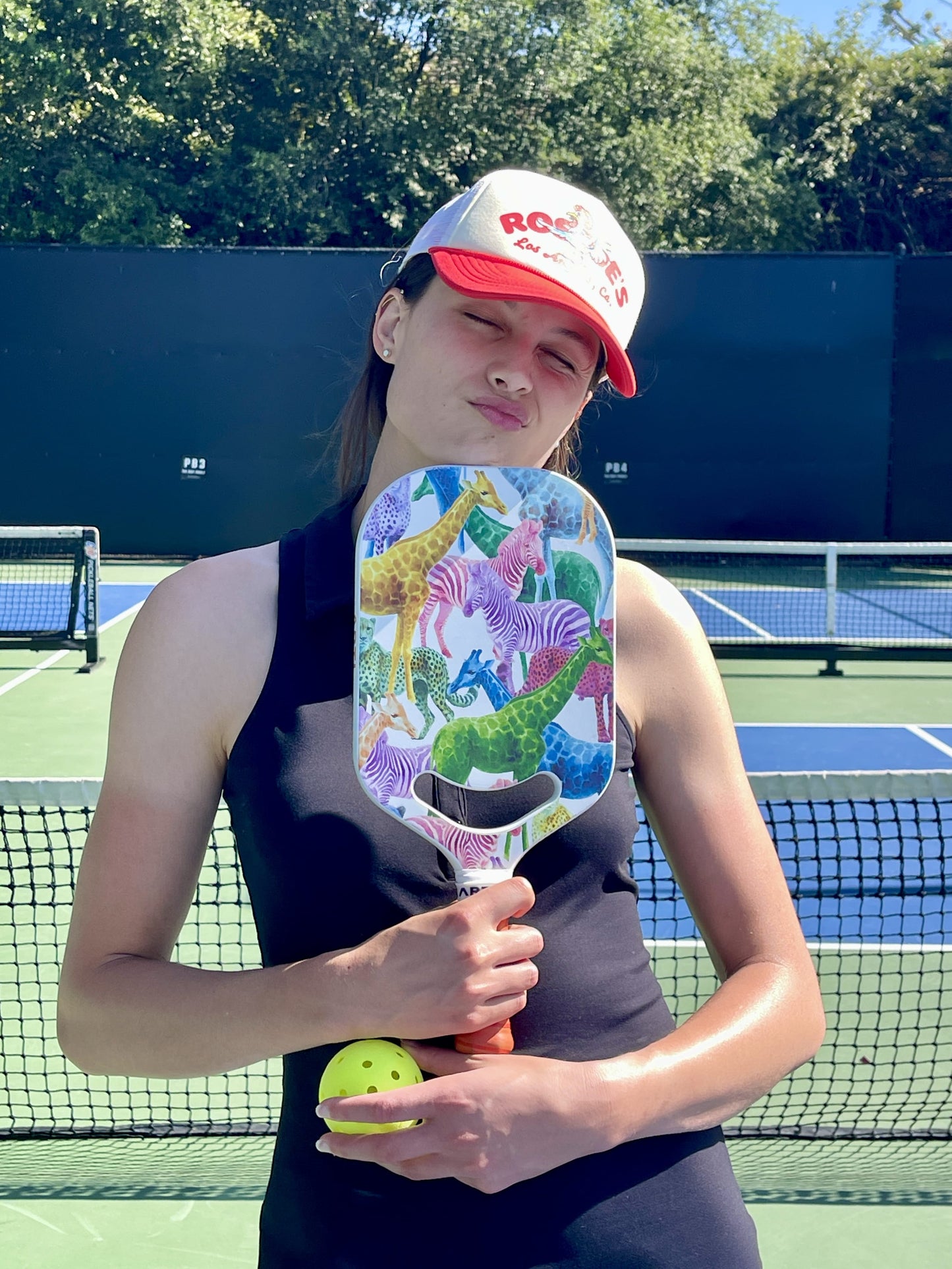 A woman in a red and white cap and black sleeveless top stands on an outdoor pickleball court, holding the ARTI Carbon Fiber EXOTIC TRIPIMAL "Animal" Paddle by ARTI. She squints in the sunlight, with trees visible in the background.