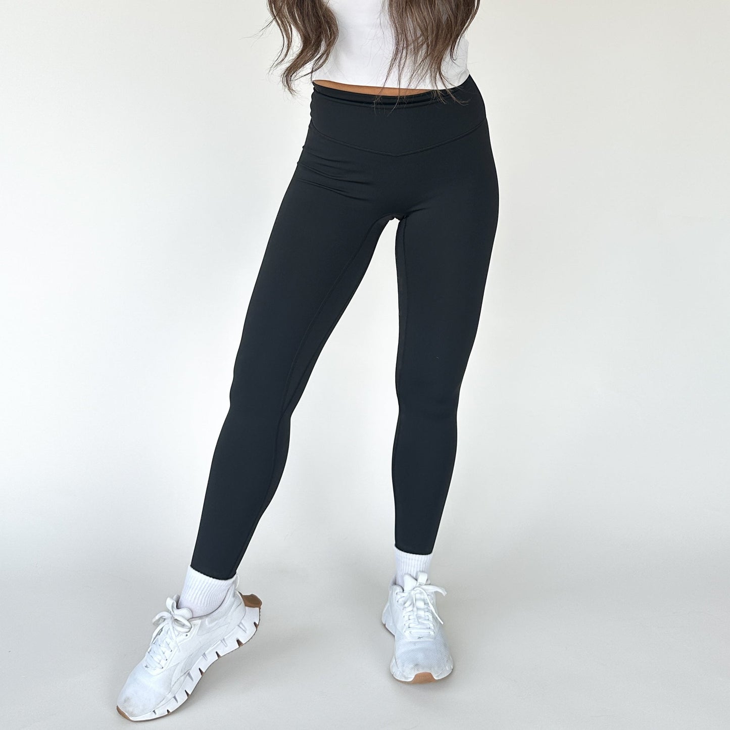 A woman showcases DYM Athletics' High Society Sculpting Legging against a white background, paired with a cropped white top, crew socks, and chunky sneakers. Her long brown hair is visible; her head is not shown.