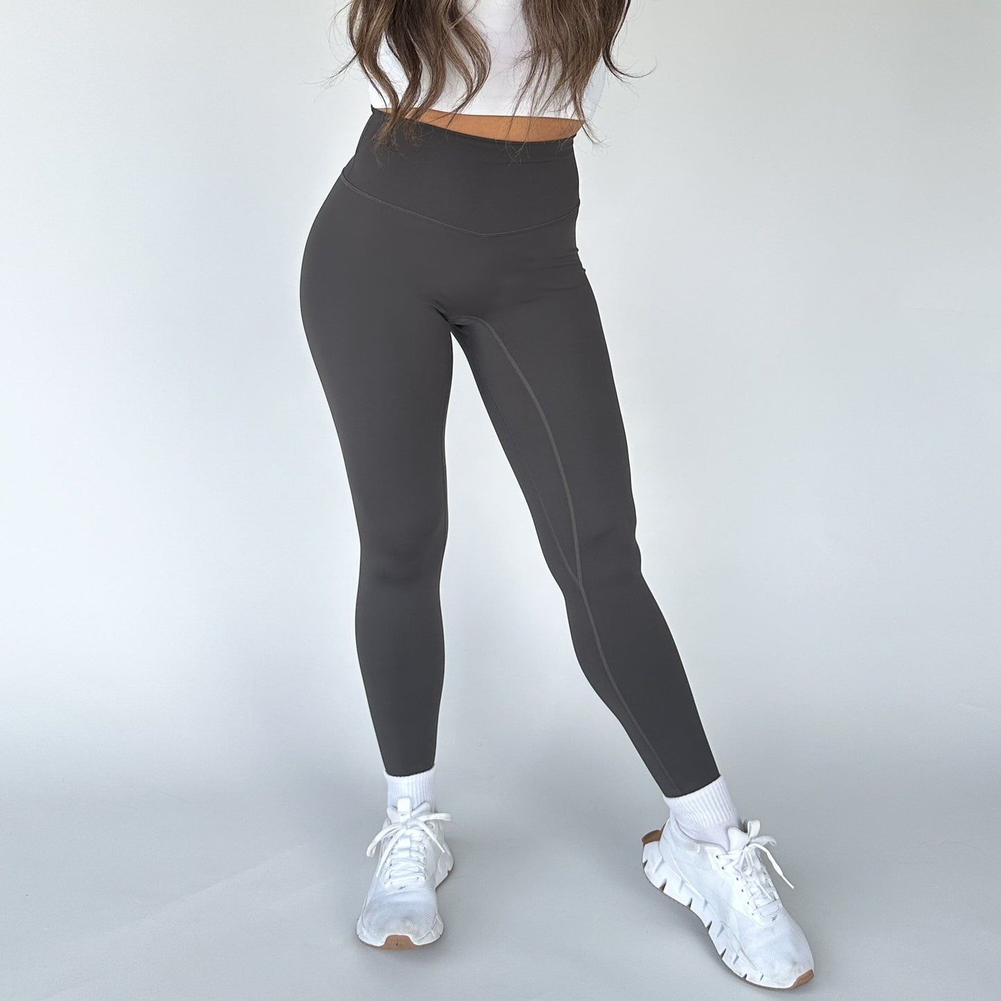 A woman models DYM Athletics' High Society Sculpting Legging in dark gray with a white crop top, white socks, and chunky sneakers. She stands against a white background; her long brown hair drapes over her shoulders, face out of frame.