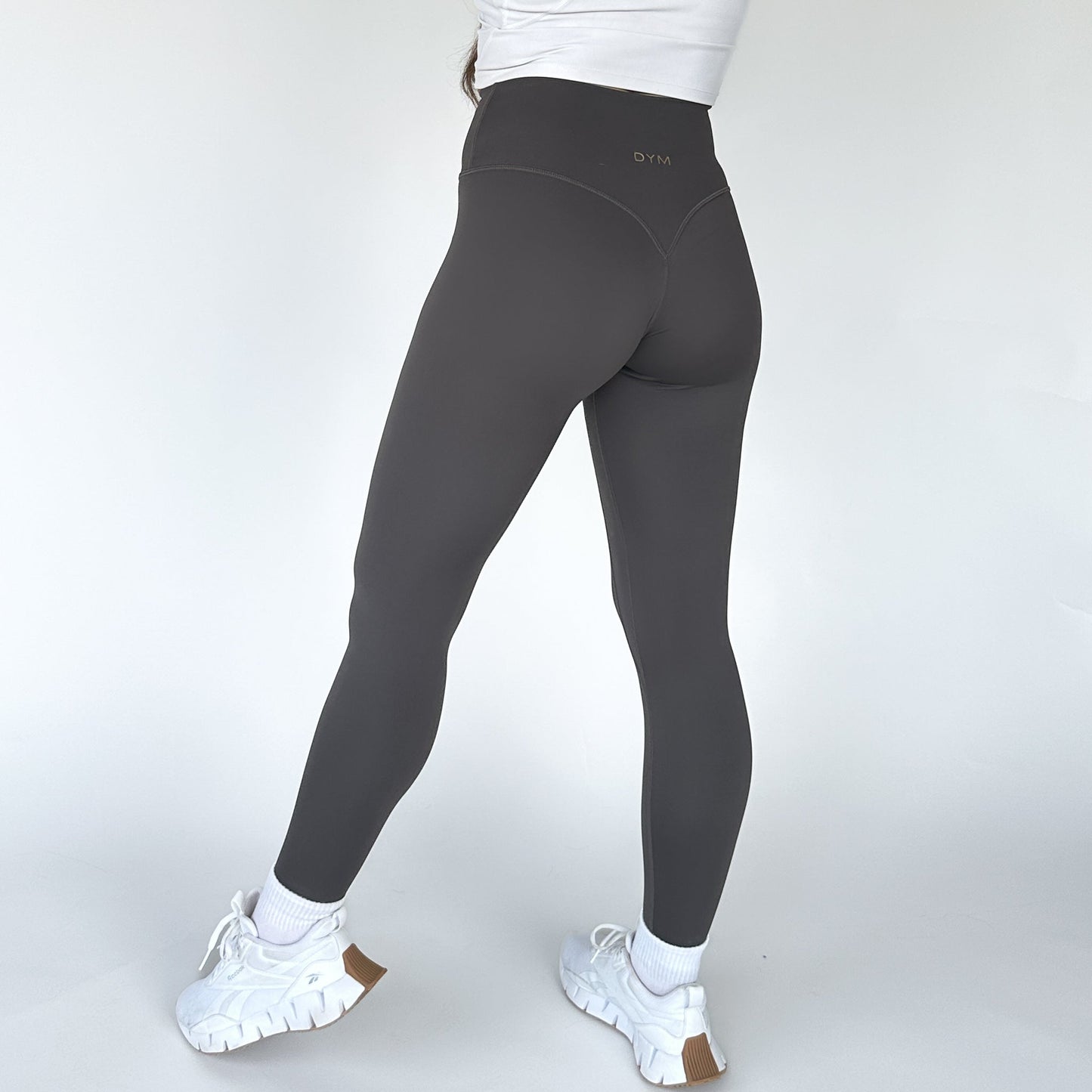 A person faces away wearing DYM Athletics’ High Society Sculpting Legging in dark gray, paired with a white crop top, white socks, and chunky sneakers. The leggings feature a small logo near the waistband against a plain white background.