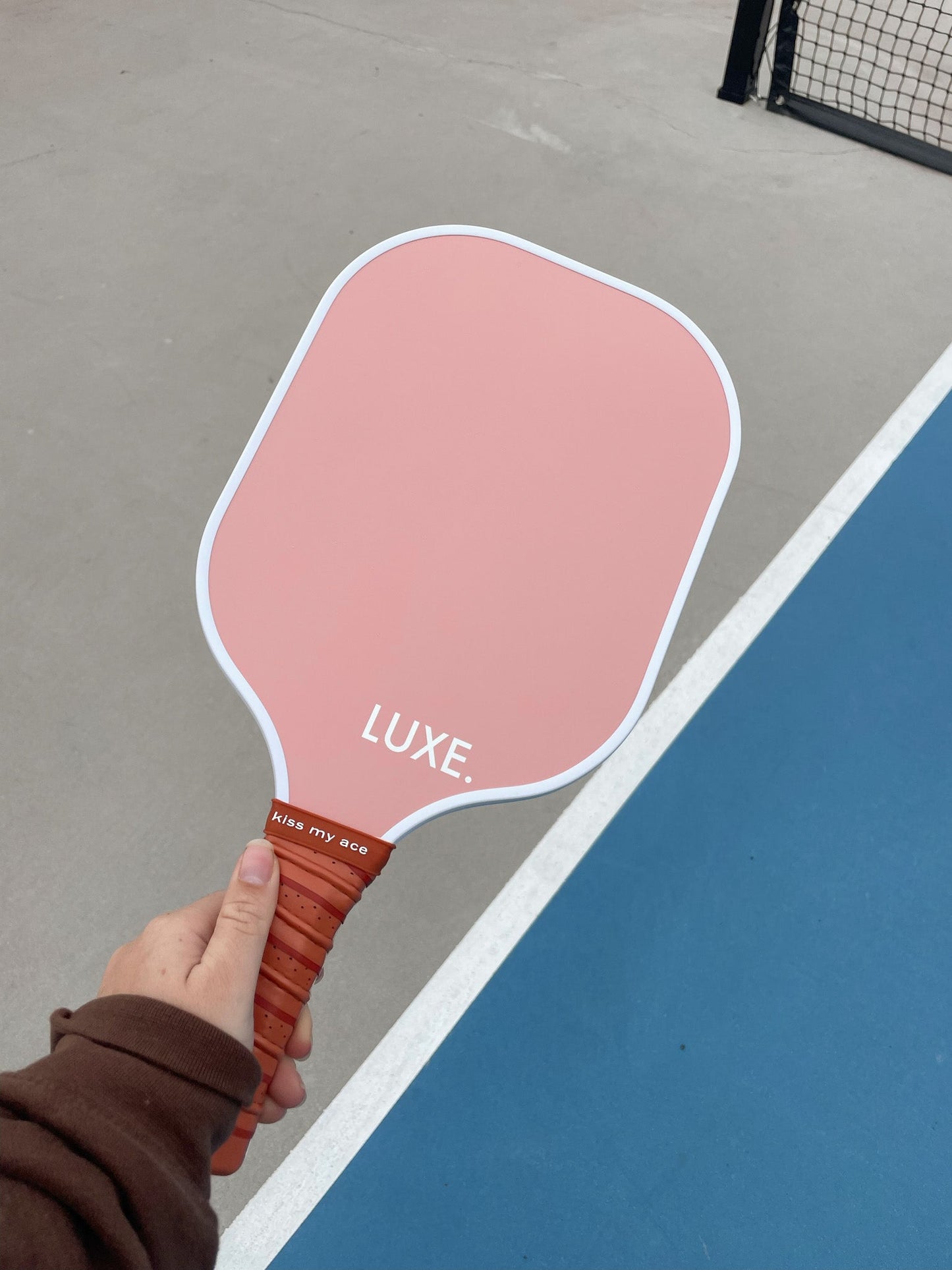 A person in a brown sleeve holds the pink Aesthetic Solids - Ace paddle by LUXE Pickleball, featuring “LUXE.” on the face. Its brown grip pops against the blue and gray court, with a black net in the background.