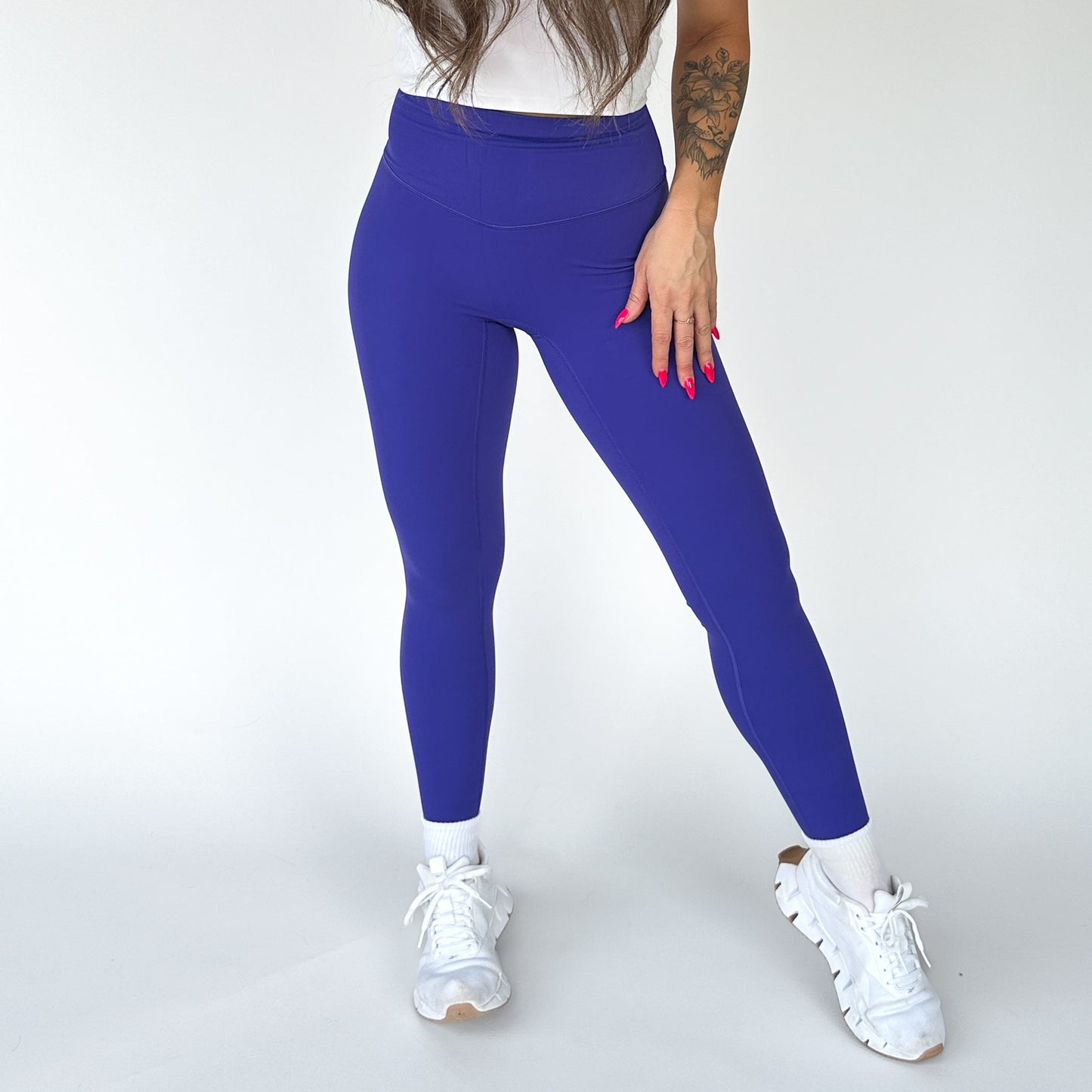 A woman wears DYM Athletics’ AIRE Ultra-Soft Legging in bright blue with tummy control, paired with a white top and sneakers. She has long hair down, a floral forearm tattoo, and bright pink nails, standing against a plain background.