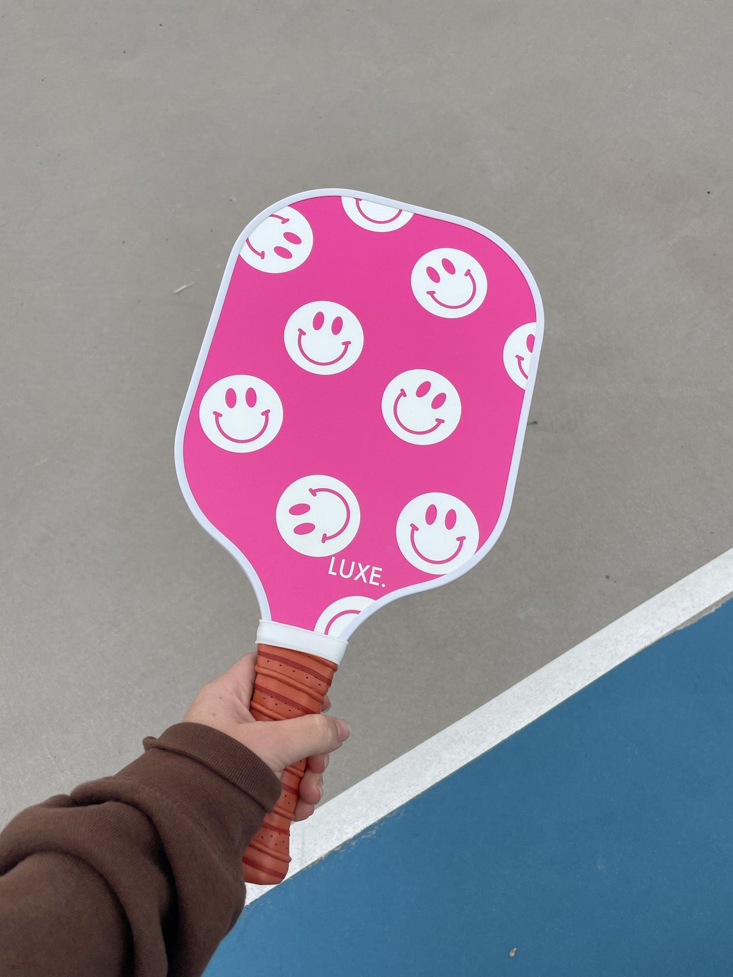 A person in a brown sleeve holds the LUXE Pickleball Smiley paddle, which is pink with white smiley faces, a fiberglass surface, brown grip, and “LUXE.” near the center, on a gray court with blue and white borders.