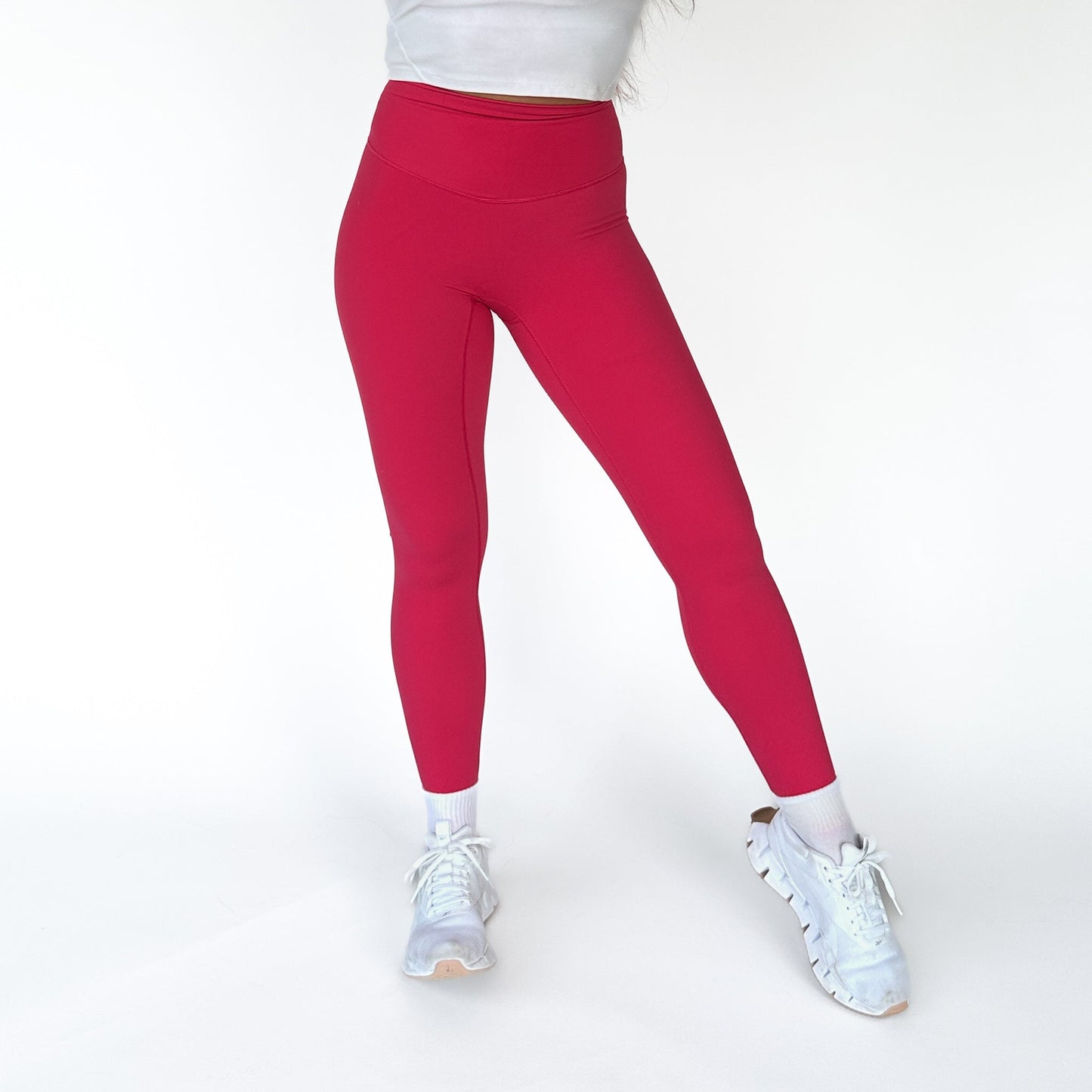 A person models the DYM Athletics AIRE Ultra-Soft Legging in bright pink, paired with a white cropped top, socks, and sneakers against a white background. Only the lower body is shown with one knee bent and feet apart.