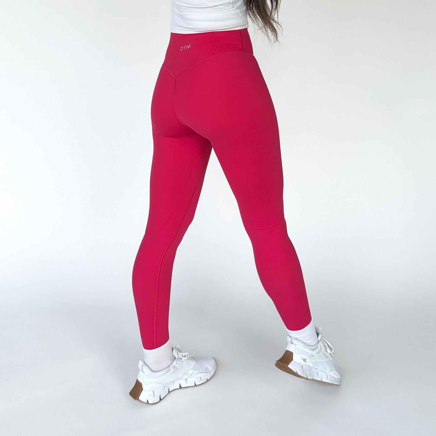A person stands with their back to the camera, wearing DYM Athletics AIRE Ultra-Soft Legging in bright pink. The high waist offers stylish support and tummy control. A small logo is near the waistband, with hair draped over one shoulder against a white background.