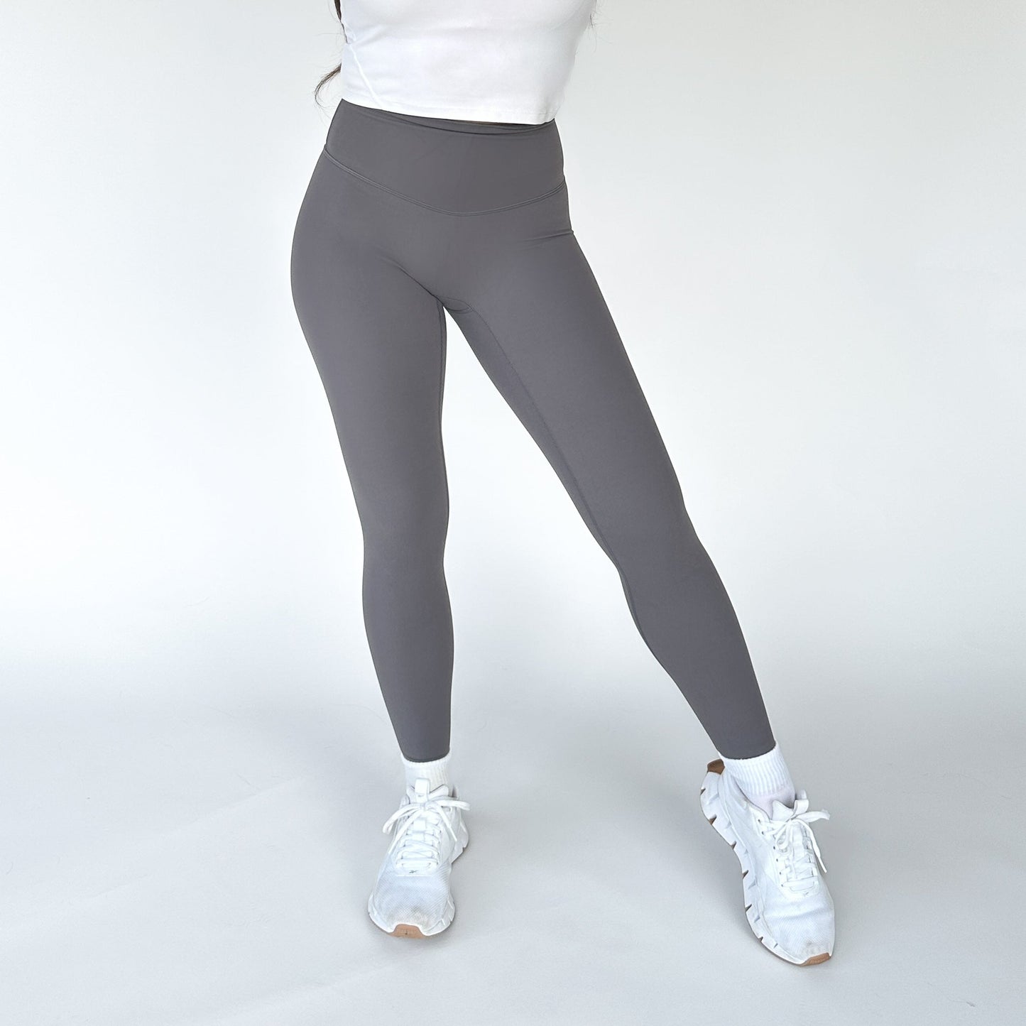 A person stands against a plain light background wearing DYM Athletics’ AIRE Ultra-Soft Legging in gray, paired with a cropped white top, white ankle socks, and athletic sneakers. Their head is out of frame; one knee is slightly bent.