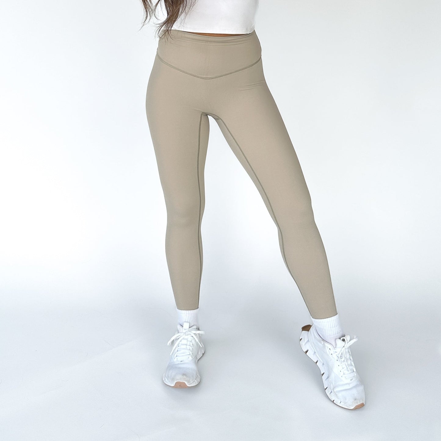 A person wears DYM Athletics’ AIRE Ultra-Soft Legging in beige, paired with a white top, white socks, and chunky sneakers. Standing on a white background, only their lower body is shown with feet apart and one knee slightly bent.
