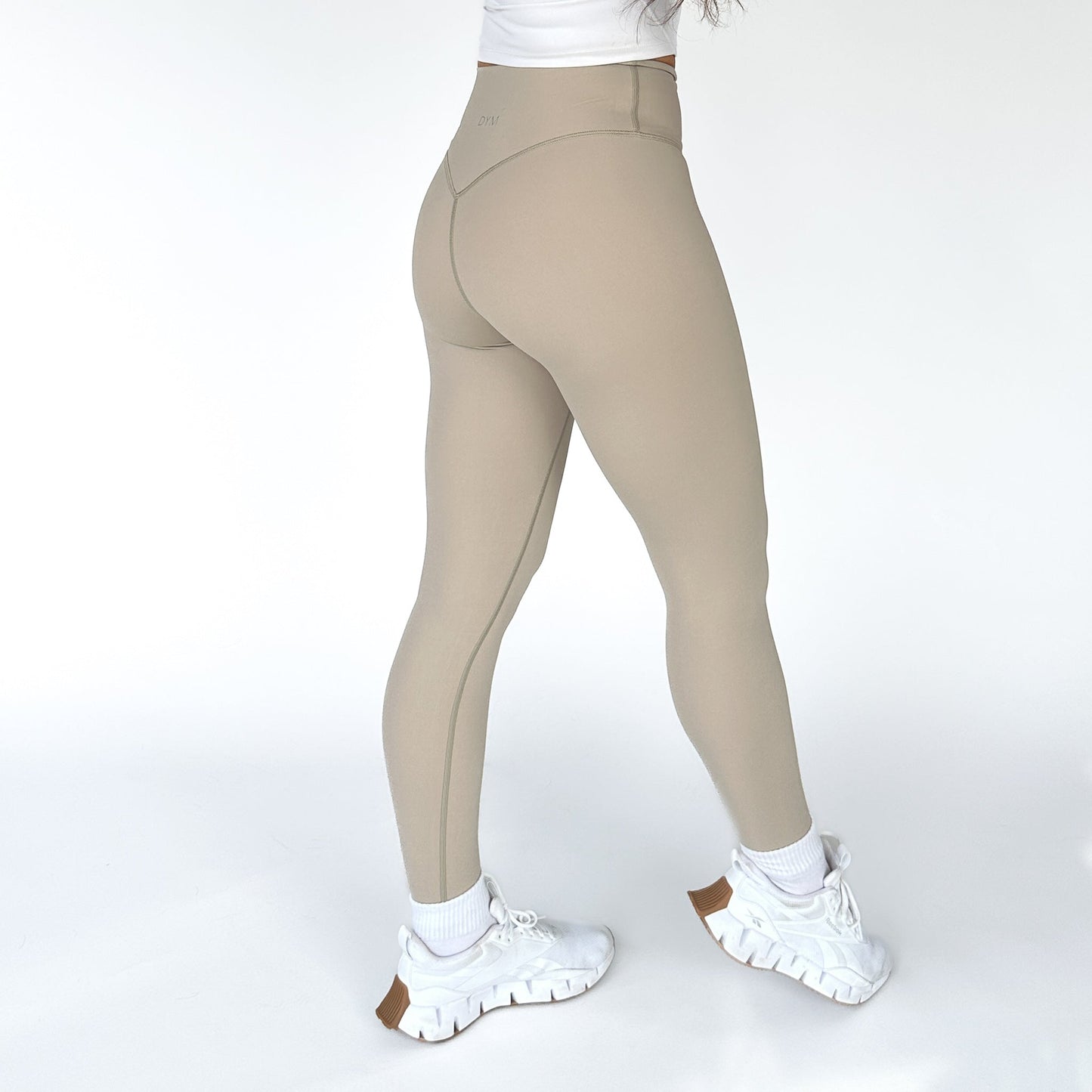 A person stands facing away, wearing DYM Athletics' AIRE Ultra-Soft Legging in beige and white chunky sneakers with brown soles. The plain white background highlights these flattering leggings and stylish footwear.