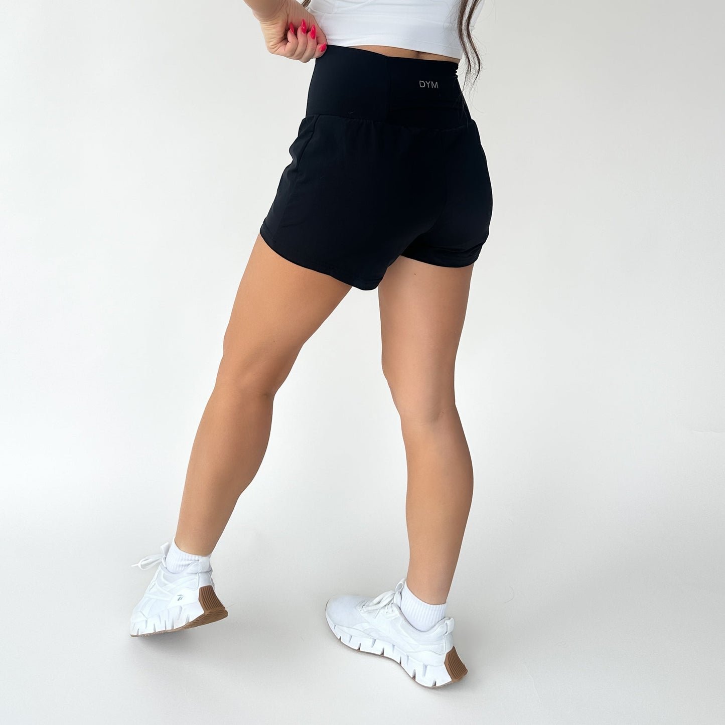 A woman faces away on a white background, wearing a white crop top, DYM Athletics Namaste Yoga Shorts | 3", white ankle socks, and chunky white sneakers with brown soles. Her left arm is bent, and her long hair falls to one side.