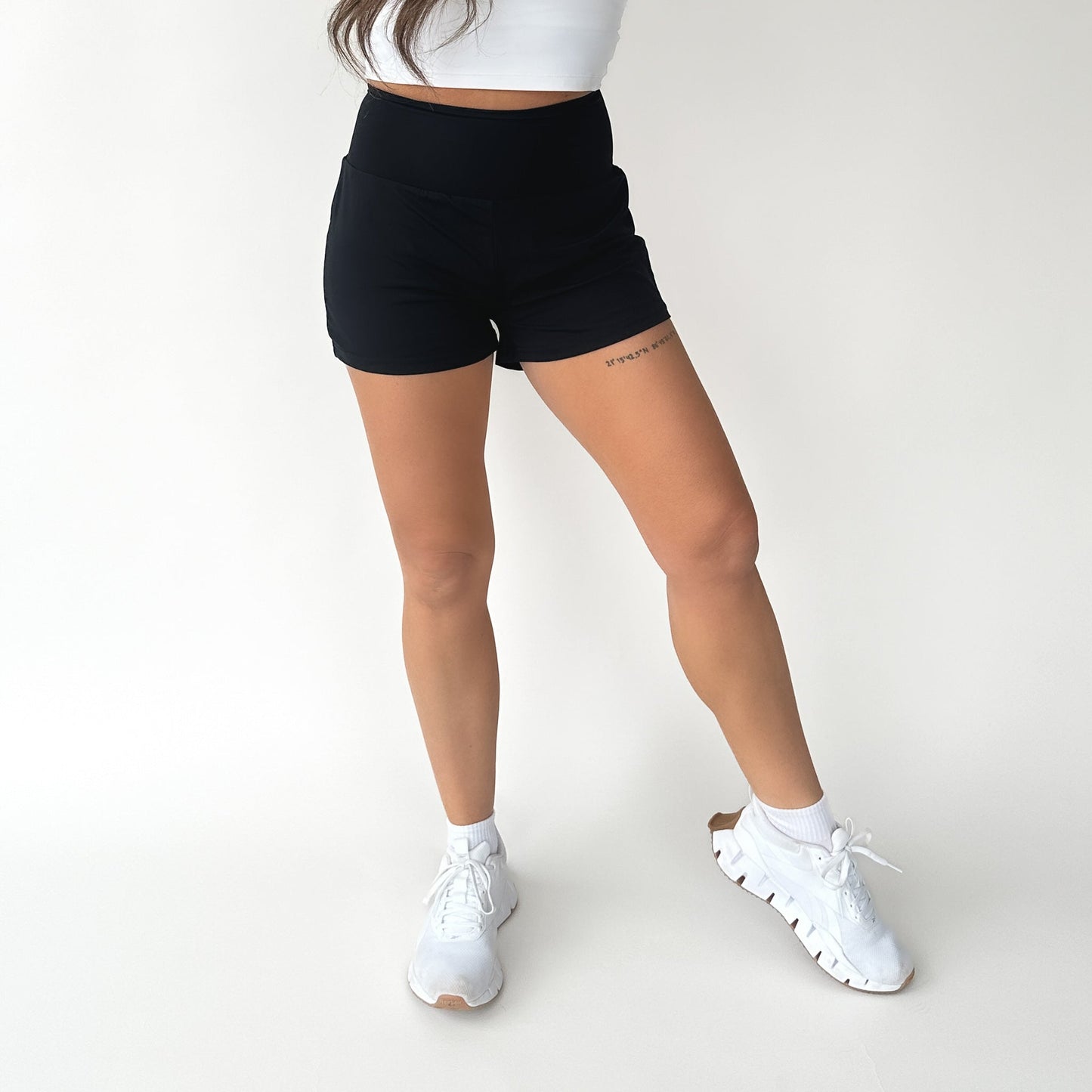 A woman poses against a white background in the DYM Athletics Namaste Yoga Shorts | 3", styled with a white crop top and athletic shoes. She stands with her long hair loose, legs slightly crossed, showing a small right thigh tattoo.