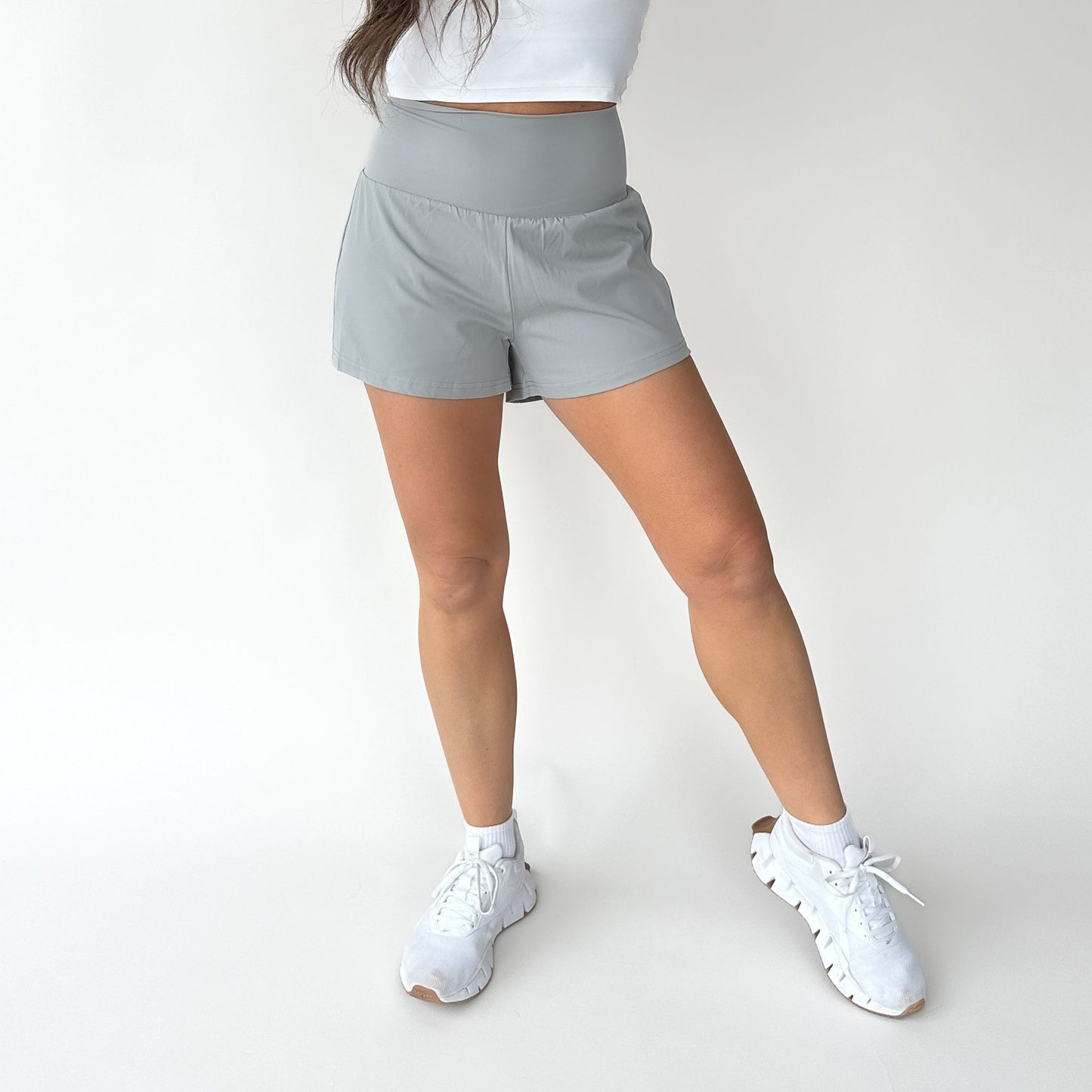 A woman, shown from shoulders down against a white background, wears a white crop top and light gray DYM Athletics Namaste Yoga Shorts | 3". Her legs are slightly apart, left knee bent, and she finishes the look with white sneakers.