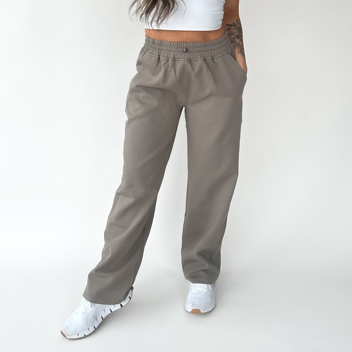 A person wears DYM Athletics Relax Wide Leg Sweatpant w/ Cinch Drawstring in taupe with a white crop top and sneakers, standing against a plain white background. Hands are in pockets, tattoo on left arm, face out of frame.