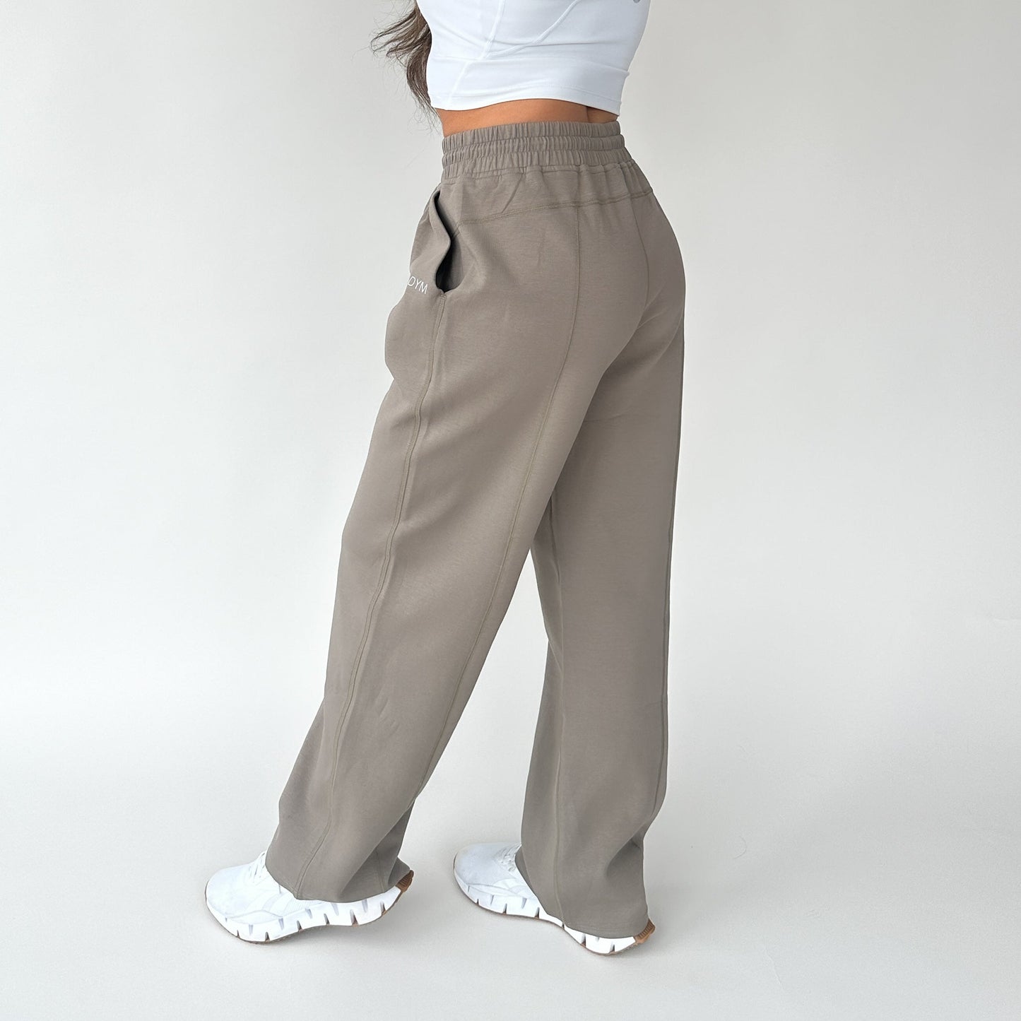 The photo shows the lower half of a person wearing DYM Athletics’ Relax Wide Leg Sweatpant w/ Cinch Drawstring in taupe, styled with a cropped white top and chunky white sneakers against a plain white background.