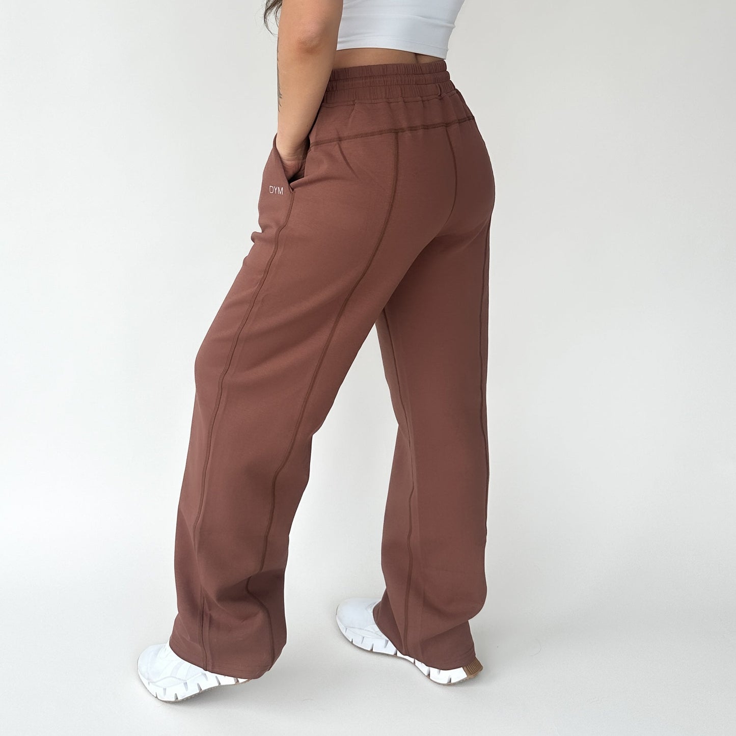 A person stands against a plain light background, wearing DYM Athletics’ Relax Wide Leg Sweatpant w/ Cinch Drawstring in brown and a white crop top. Their left hand is in their pocket, wearing white sneakers. Their head and face are not visible.