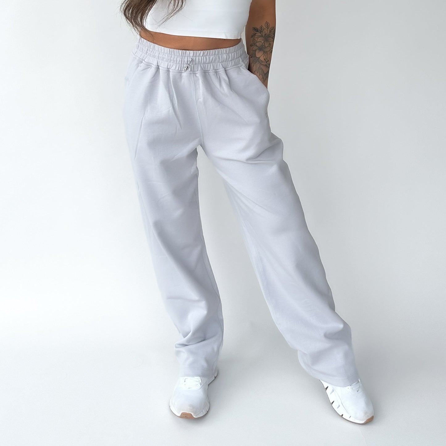 A person stands against a white background wearing DYM Athletics’ Relax Wide Leg Sweatpant w/ Cinch Drawstring in light gray, paired with a white cropped top and white sneakers. Only the shoulders down are visible; one arm has a tattoo. Hands are in pockets.