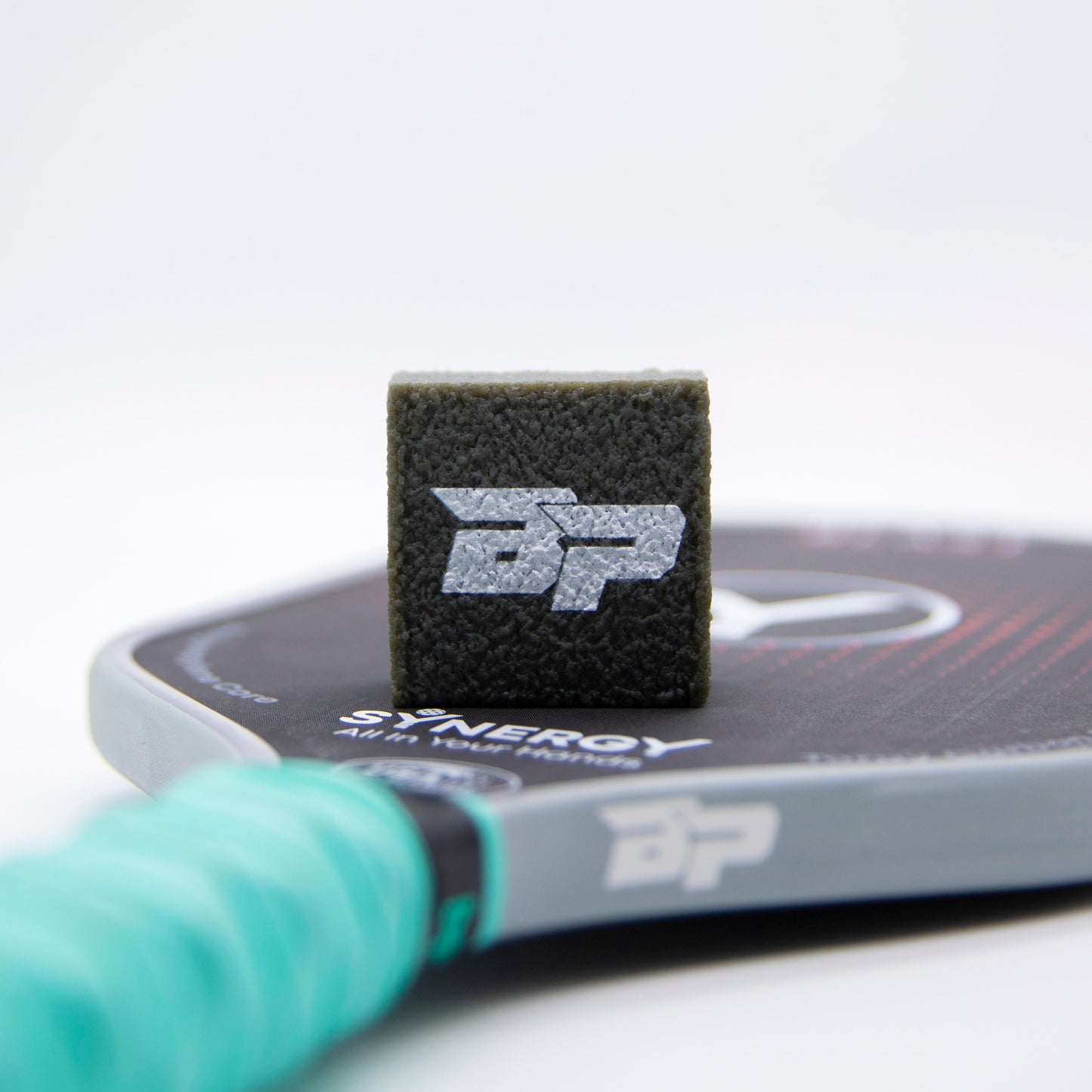 Close-up of a Bodhi Performance SYNERGY Air-Tek Power pickleball paddle with a black edge guard and textured white “BP” logo. A PROeraser™ cleaner rests on the paddle, which has a turquoise grip and white branding, set against a white background.