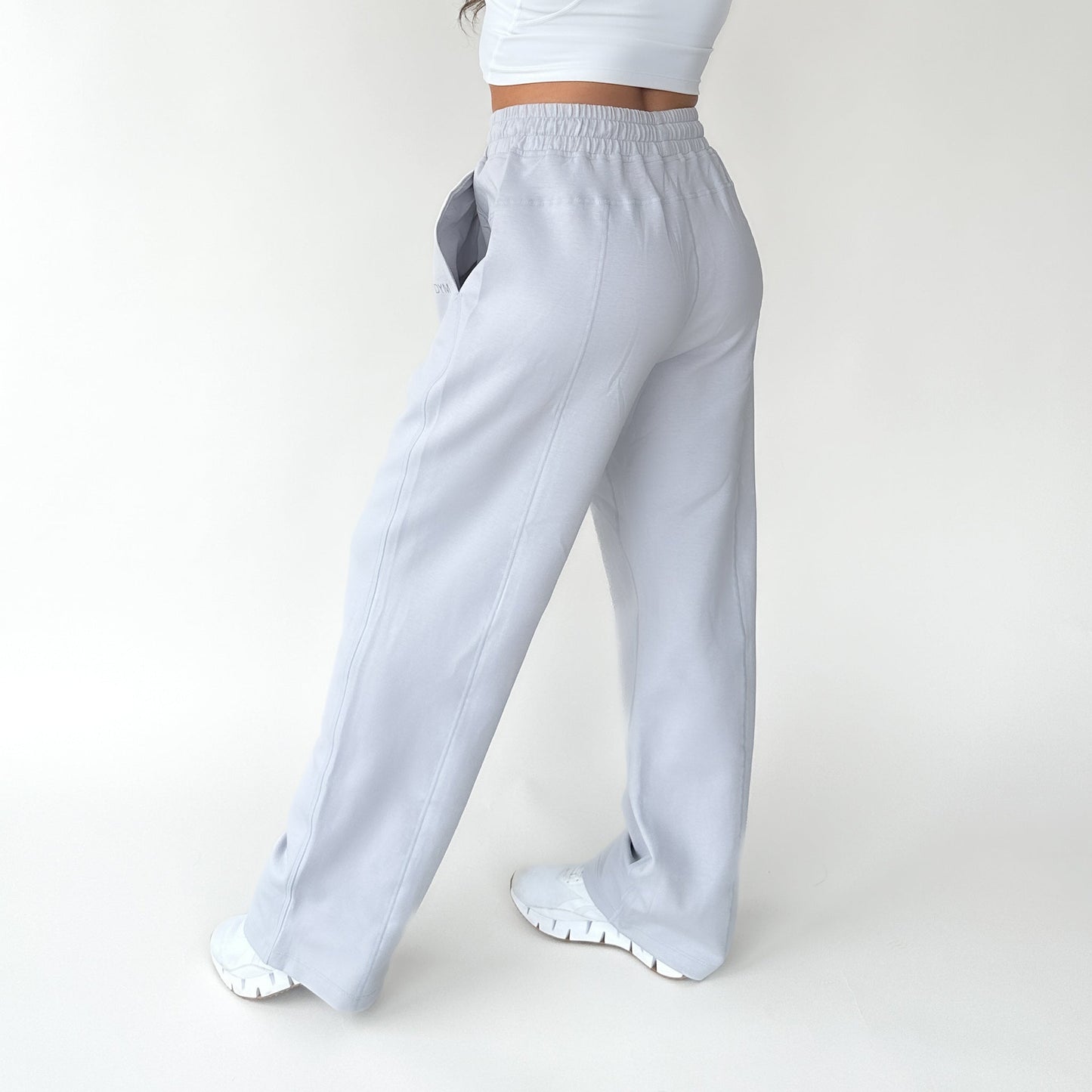 A person wears DYM Athletics Relax Wide Leg Sweatpant w/ Cinch Drawstring in light gray with white sneakers and a white top, standing against a plain white background. The back and side views of the cozy loungewear pants are clearly visible.