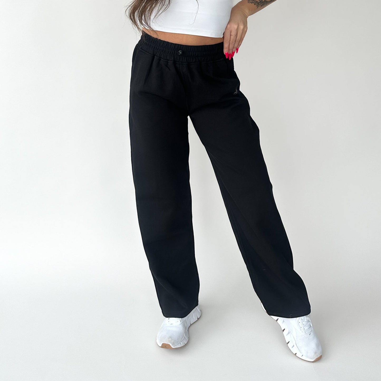 Against a plain white background, a person models the DYM Athletics Relax Wide Leg Sweatpant w/ Cinch Drawstring with a cropped white top and white sneakers. Their hand, sporting bright pink nails and a forearm tattoo, rests on their hip; face not shown.